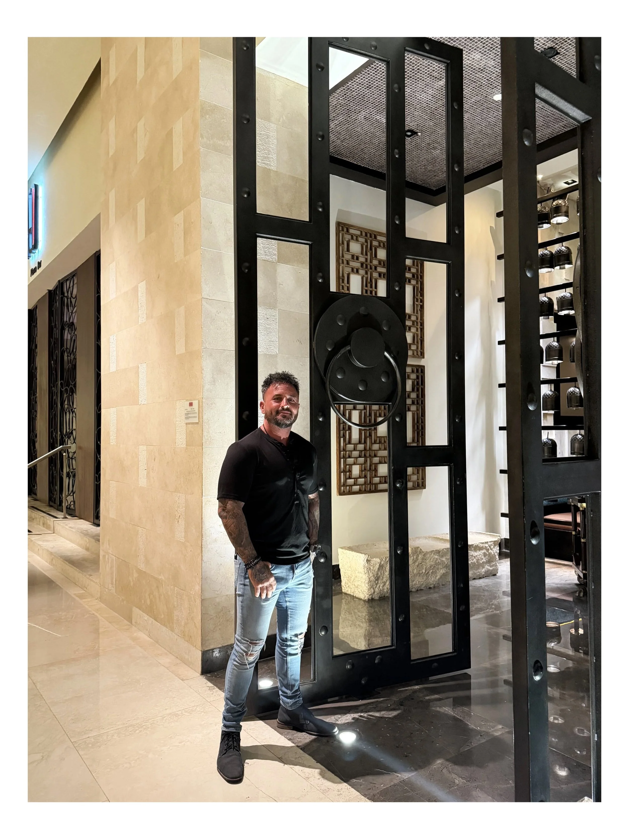 A man with tattoos on his arms, wearing a black shirt, distressed jeans, and black shoes, standing indoors near a large black metallic structure with geometric rectangular openings and circular elements. The background features beige tiled walls, decorative wall art, and shelves with black objects, in a modern interior setting.