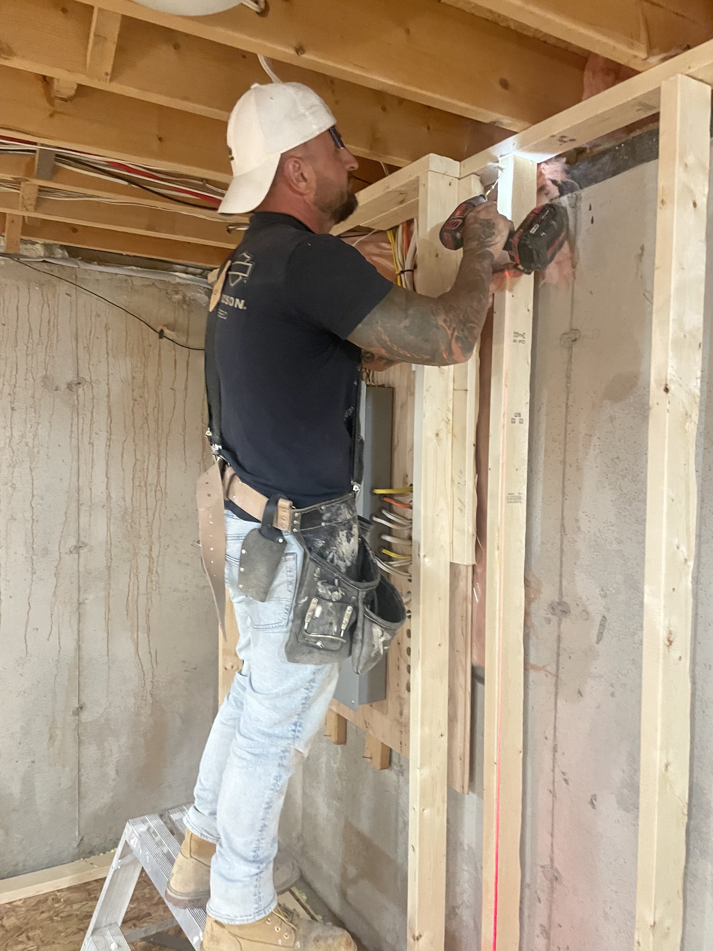 A man wearing a white cap, black T-shirt, jeans, and work boots standing on a small ladder working with a cordless drill on a wooden frame at a construction site. He has tattoos on his left arm and a tool pouch attached to his waist.