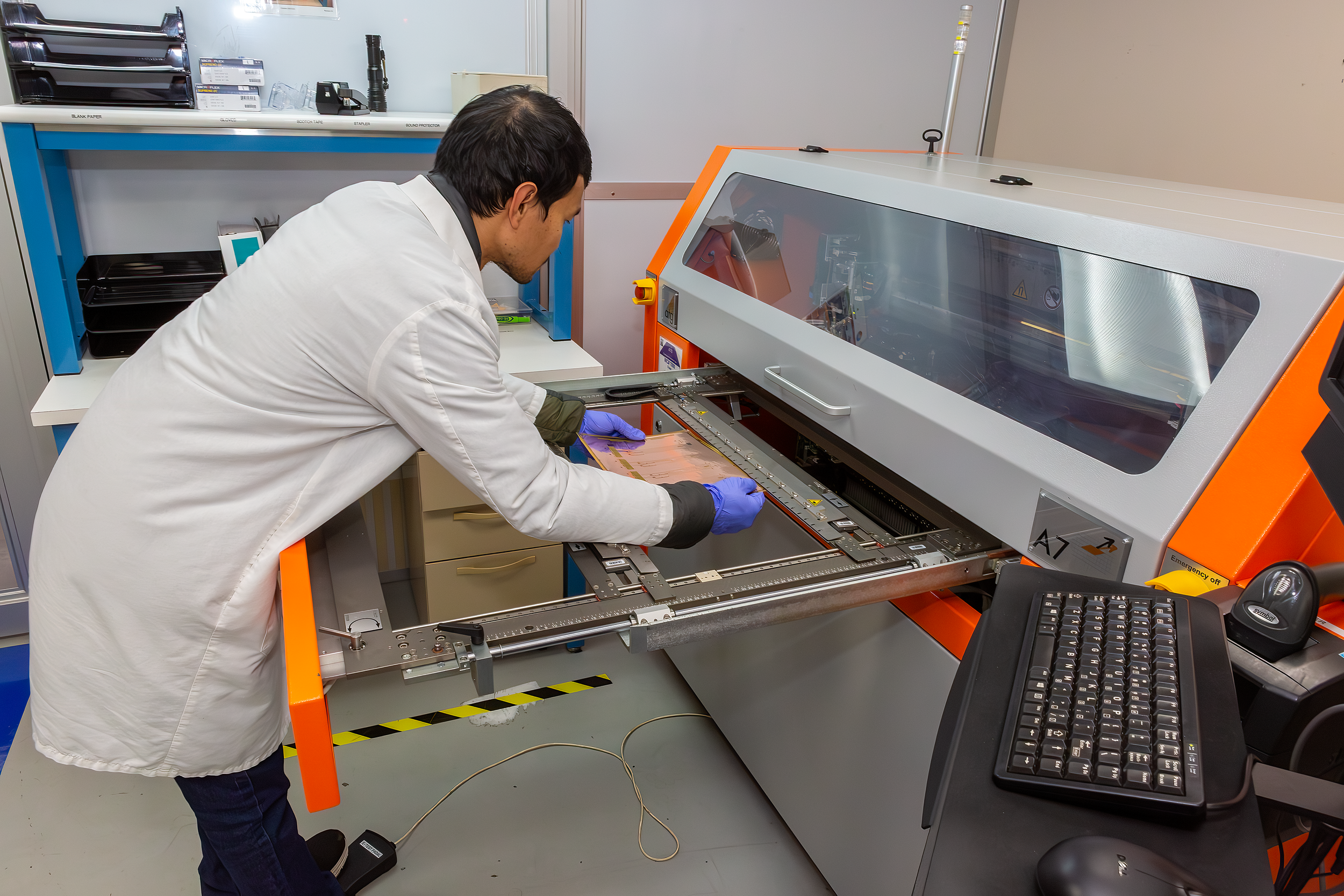 A technician wearing a white lab coat and purple gloves working on a large experimental instrument in a laboratory. He is bent over with his head close to the machine, which has a transparent cover and orange framing. The lab has shelves with various supplies, a keyboard, and a computer mouse on a desk.
