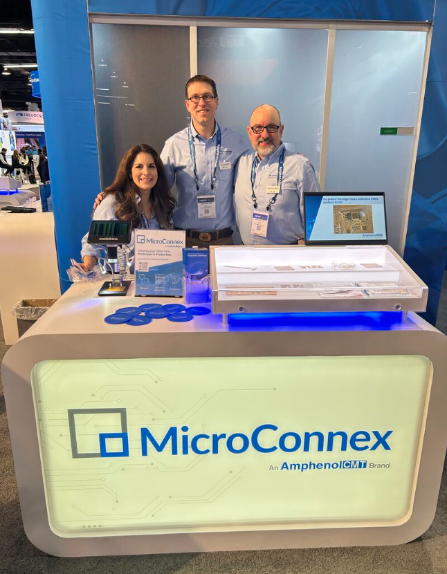 Three people at a MicroConnex booth at a trade show or conference, standing behind a display counter with promotional materials and circuit prototypes, with the MicroConnex logo on the front of the counter.