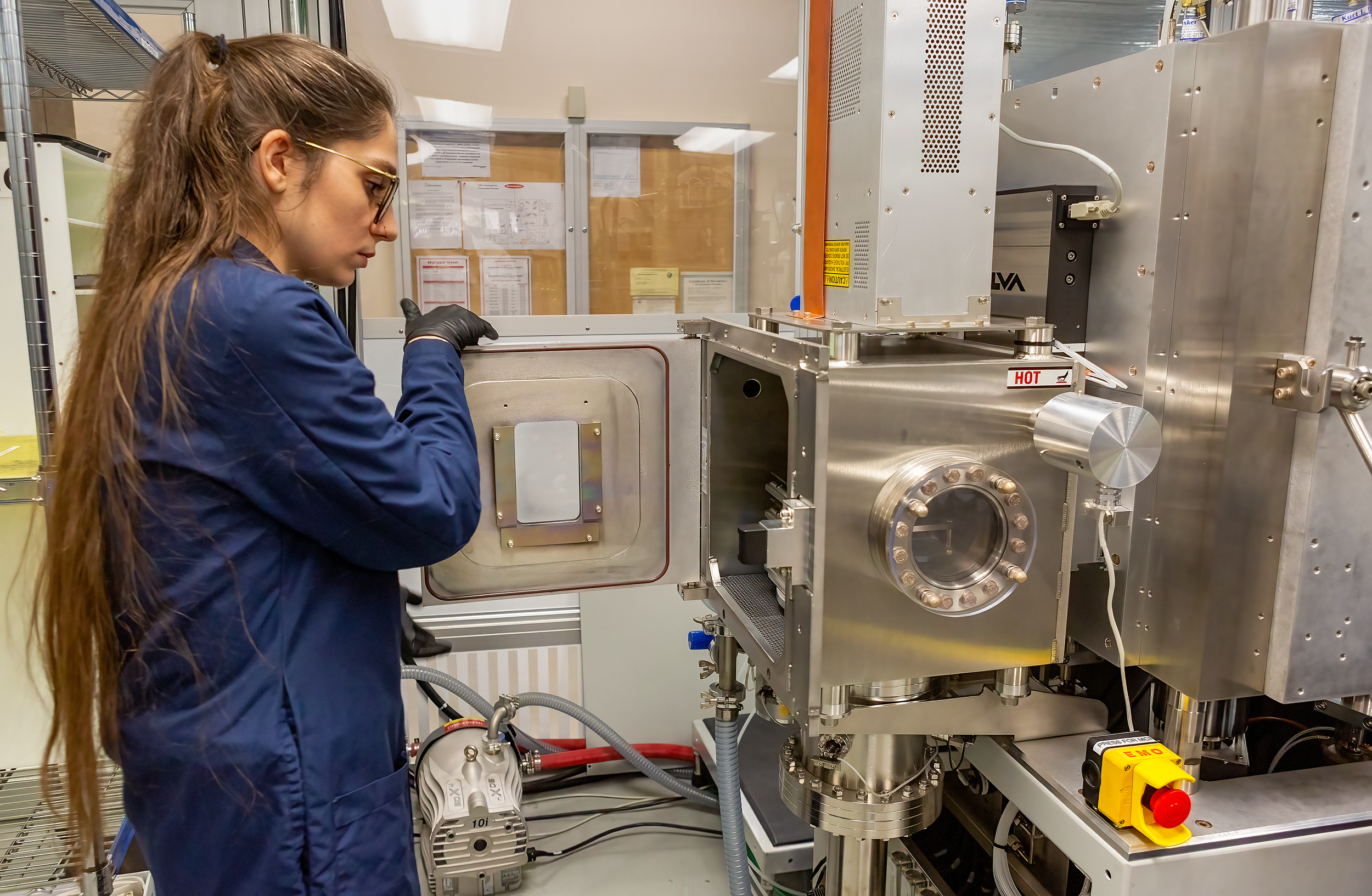 A woman in safety glasses and a blue lab coat working with a large scientific instrument in a laboratory.