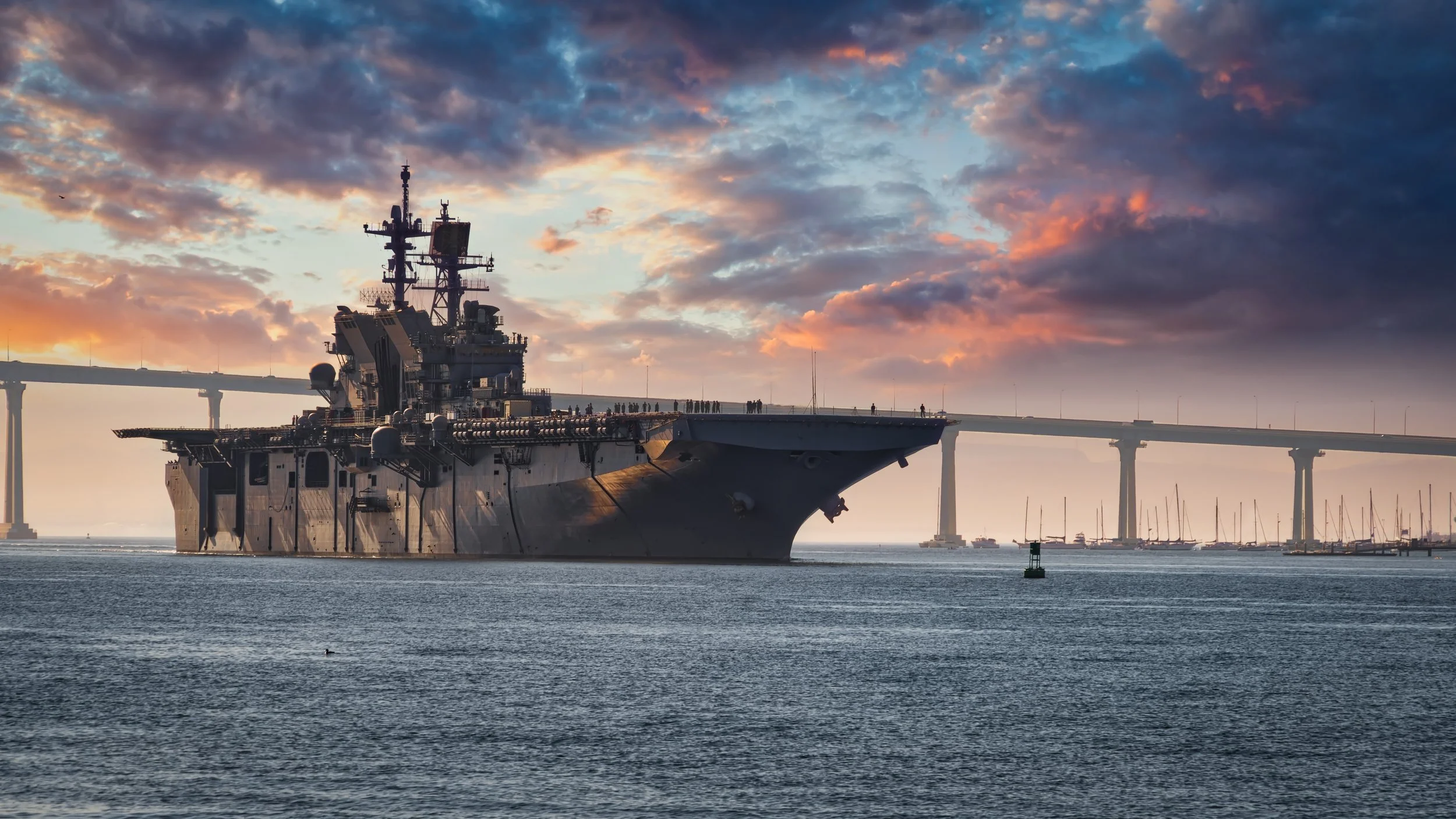 A large aircraft carrier ship sailing in calm waters during sunset with a bridge in the background and a cloudy sky with orange and purple hues.