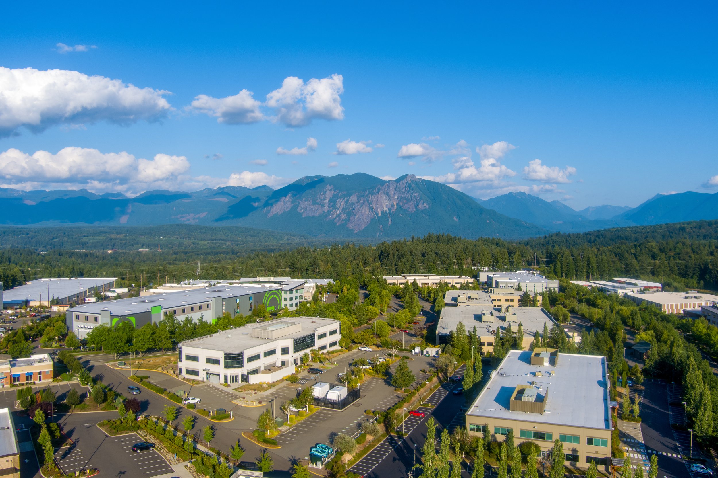 A cityscape with commercial buildings, green parking lot, trees, and mountains in the background under a blue sky with clouds.