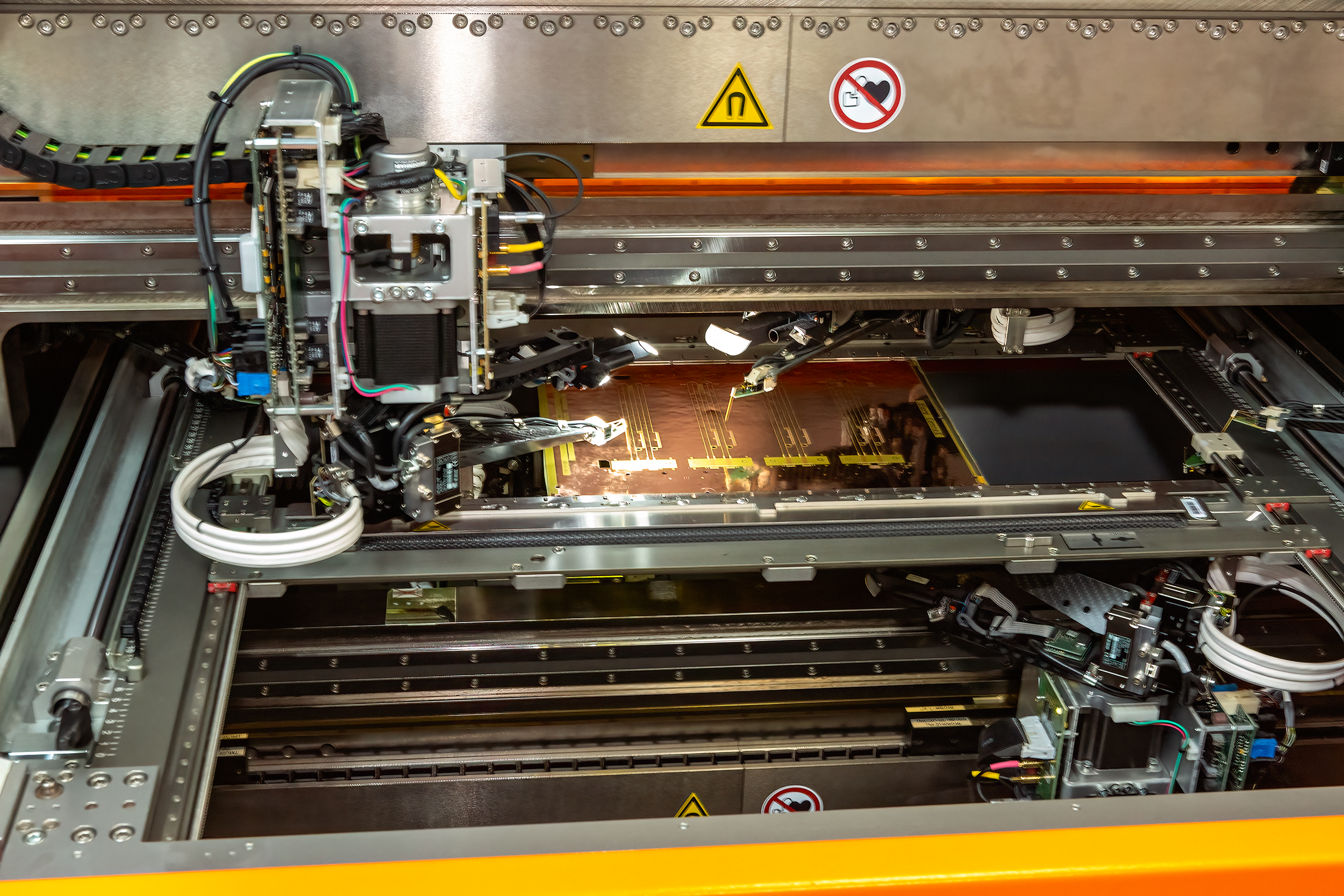 The image shows the inside of a machine or industrial equipment with various electronic components, wiring, and warning labels, including a yellow caution sign and a no-hand-in-moving-parts sign.