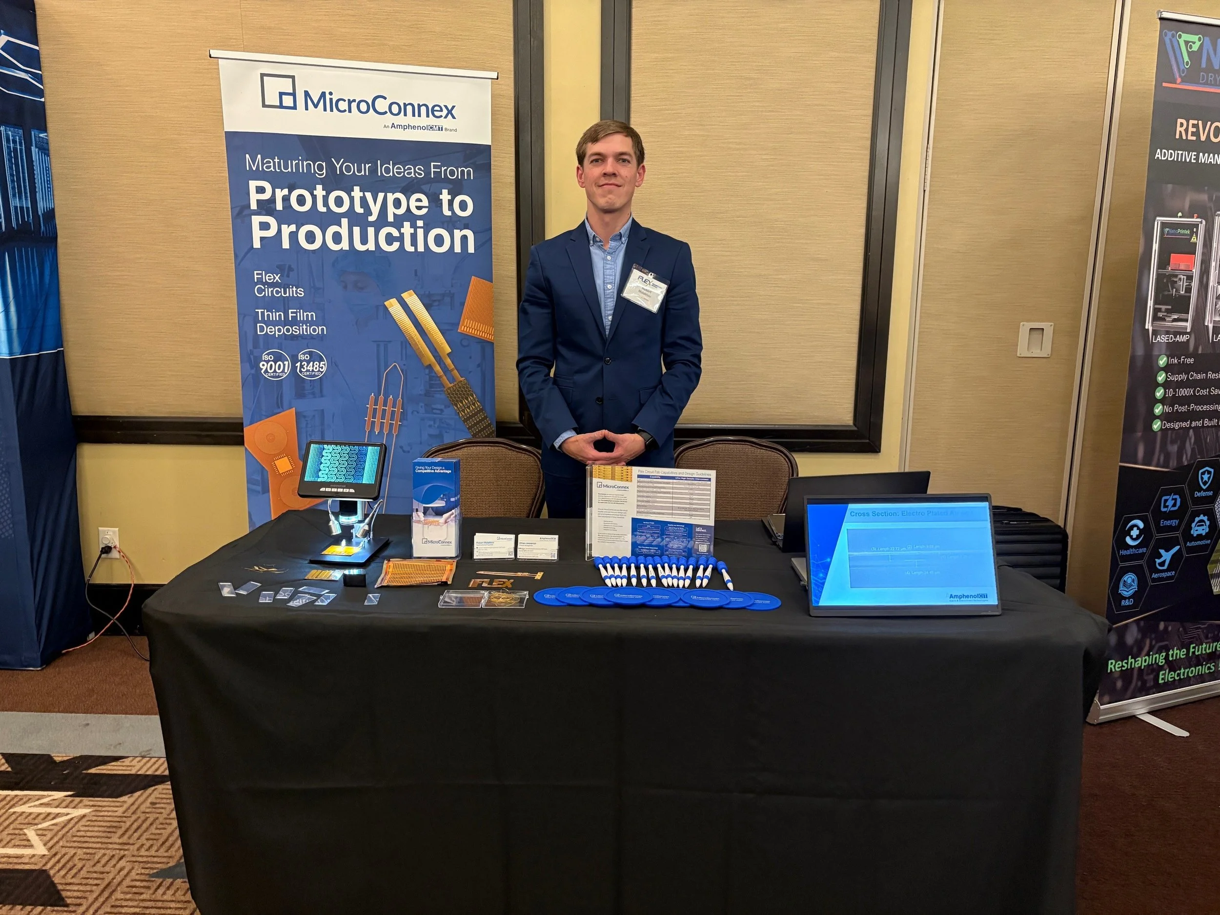 A man in a blue suit standing behind a table at a conference booth with a MicroConnex banner that reads "Maturing Your Ideas From Prototype to Production," with images of electronic components and 3D printed materials displayed on the banner and table.