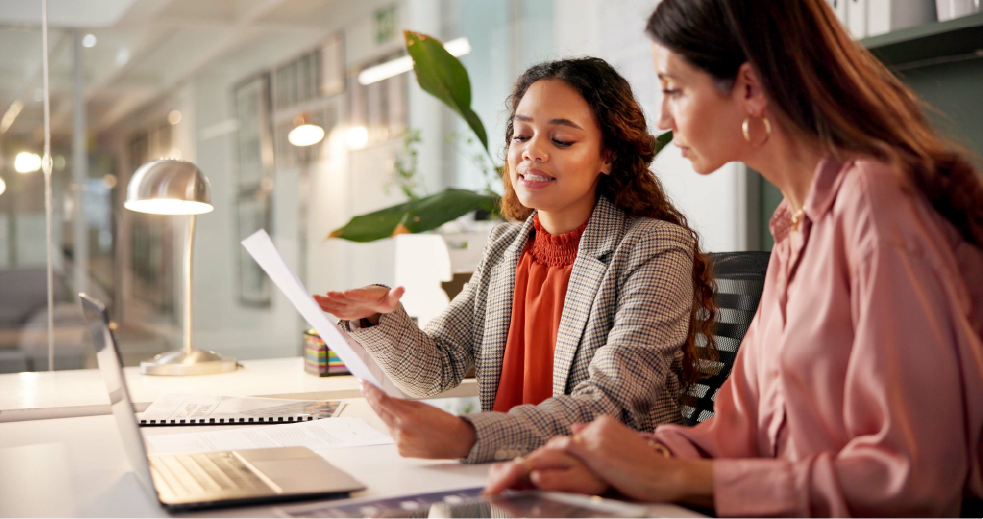 Two women sitting at a desk in a modern office, looking at a document together, with a laptop open in front of them. One woman has curly hair and is wearing a checked blazer, while the other has straight hair and is wearing a pink blouse.