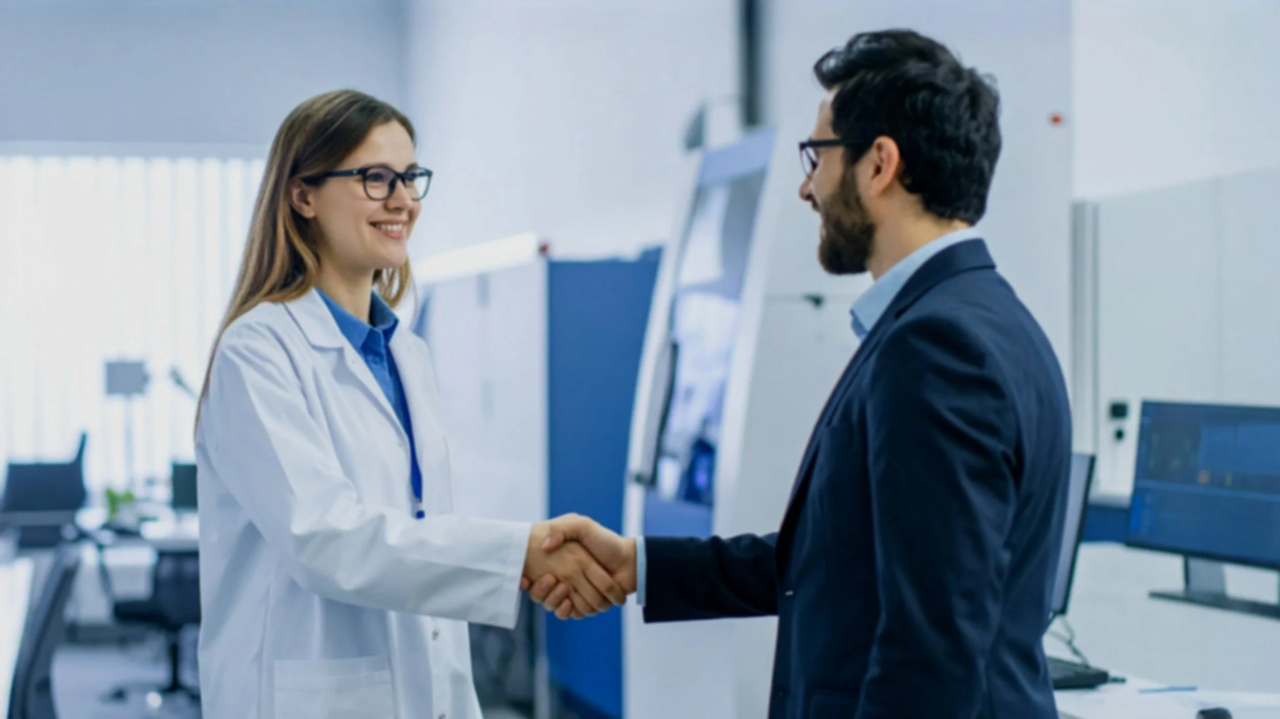 A woman in a white lab coat shaking hands with a man in a suit in a modern laboratory.