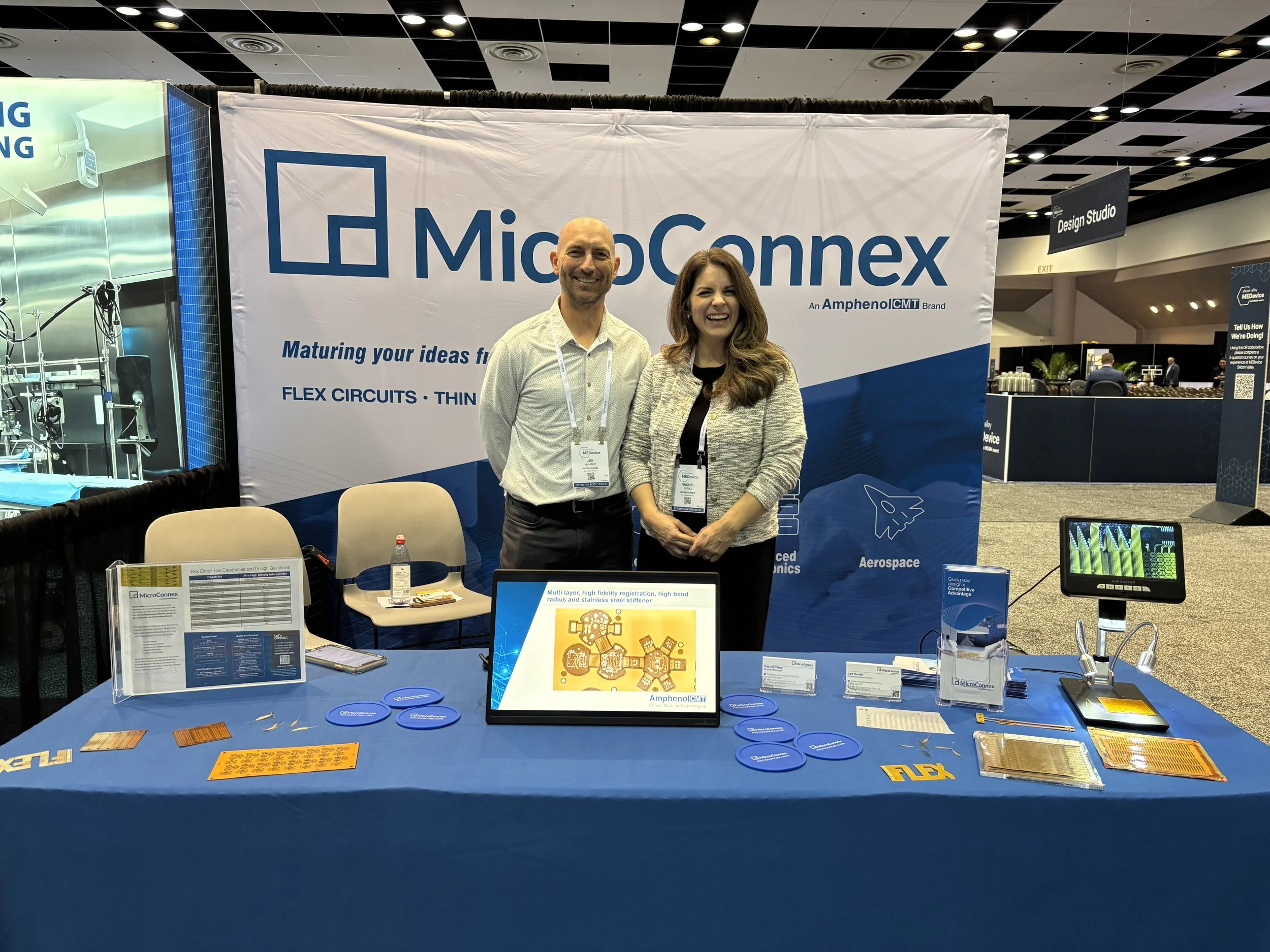 Two people stand behind a booth at a trade show with a large banner displaying the MicroConnex logo and slogan. The booth has electronic components and informational materials on the table in front of them.