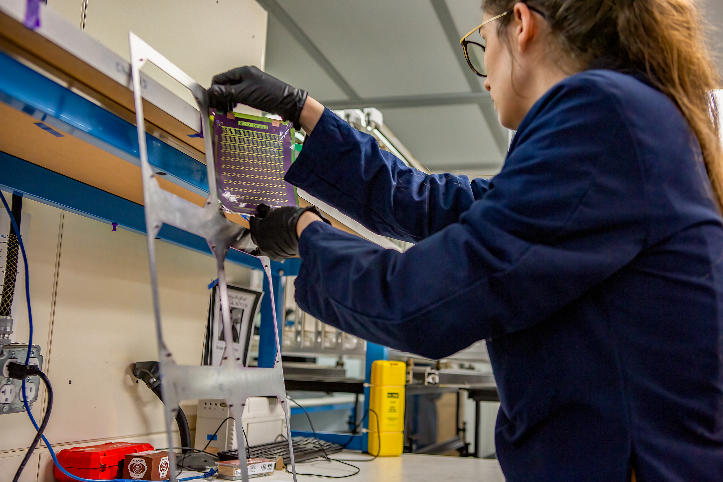 A woman working in a laboratory, placing or adjusting a circuit board on a rack. She is wearing glasses and black gloves, with various electronic equipment and tools around her.