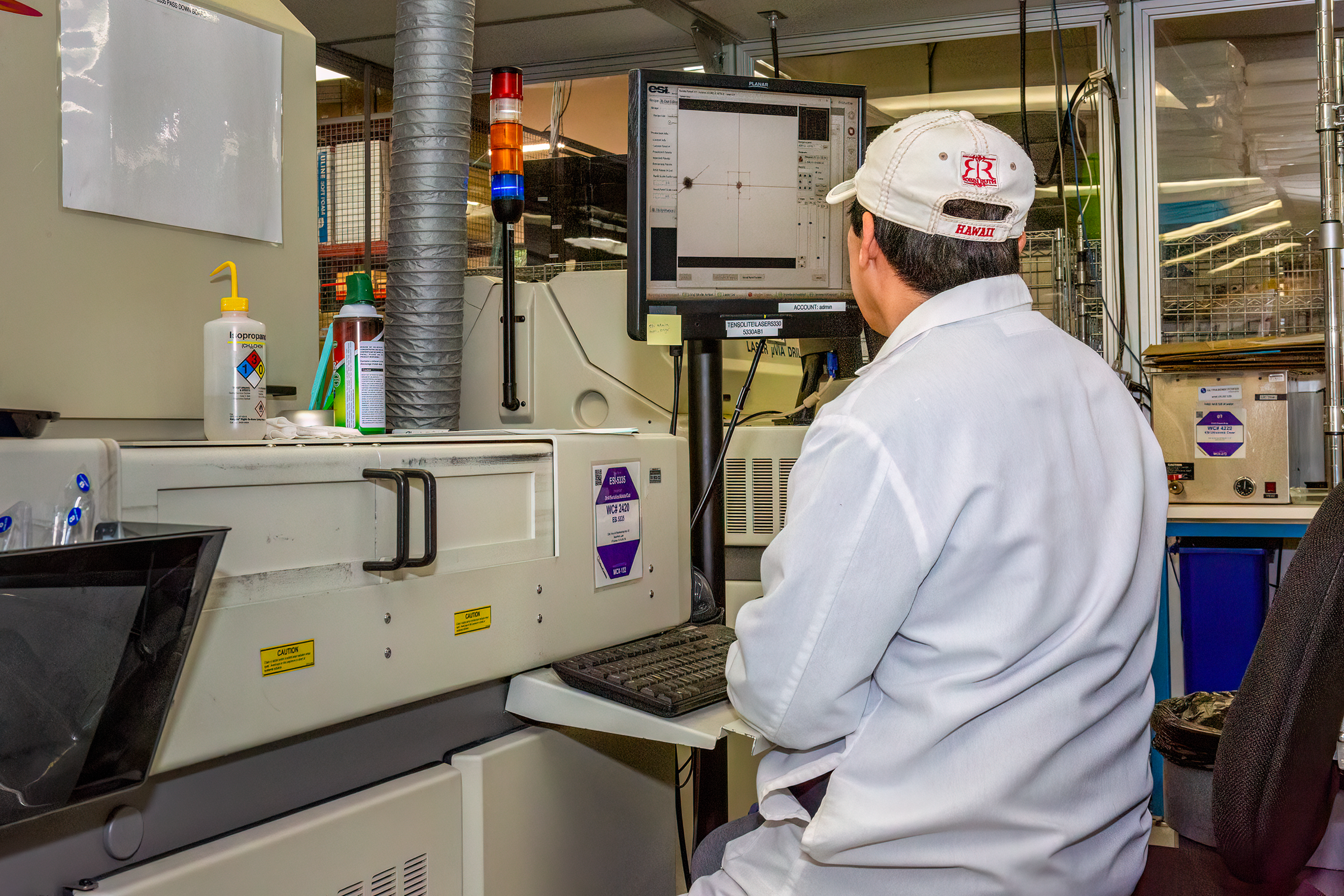 A person wearing a white lab coat and a white cap with red lettering working at a laboratory instrument in a lab environment. There are chemicals and lab equipment on the counter, and a large monitor displaying data.