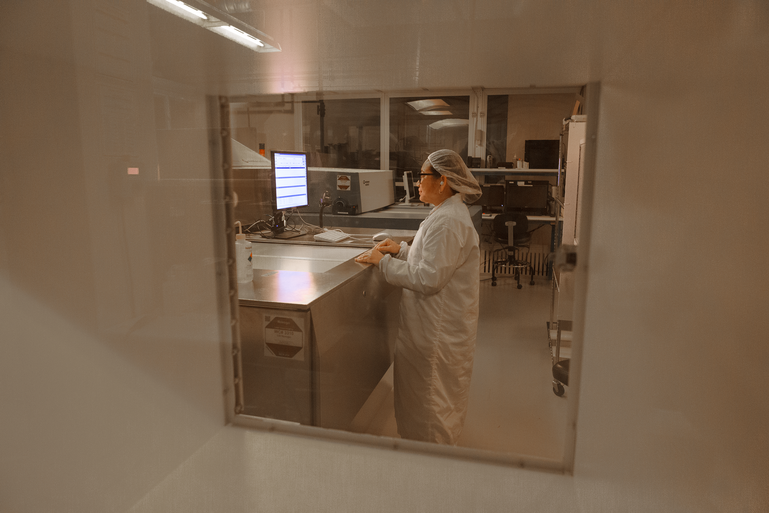A person wearing a lab coat, hairnet, and glasses working at a computer in a laboratory or cleanroom, viewed through a window.