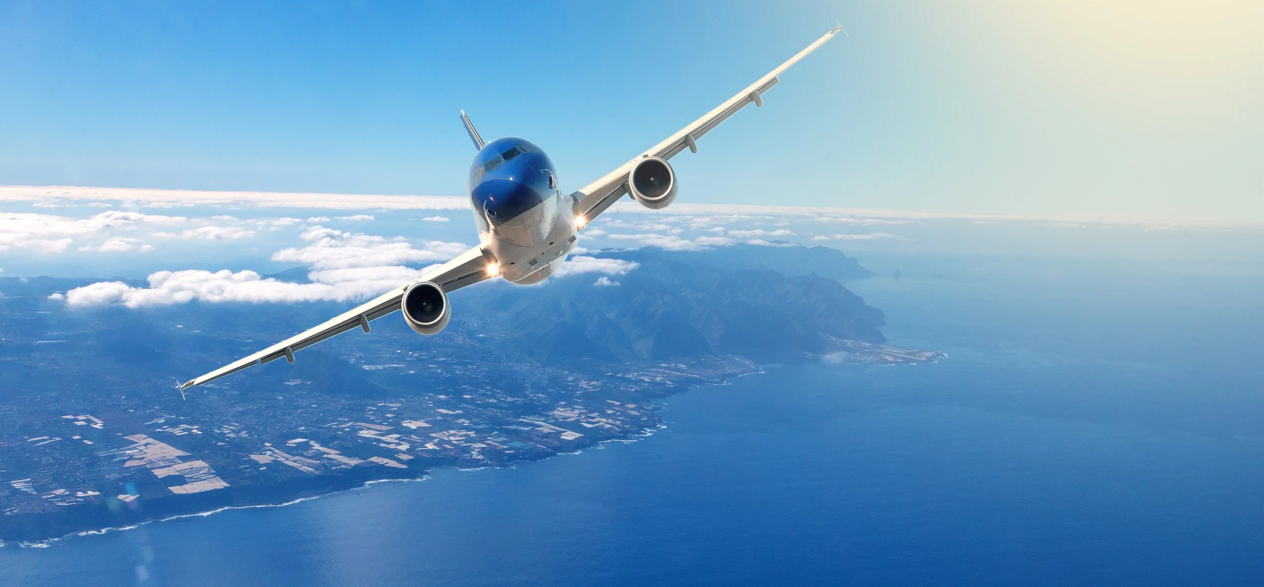 A commercial airplane flying high above a landscape with mountains, islands, and water, with a clear blue sky and some clouds around.