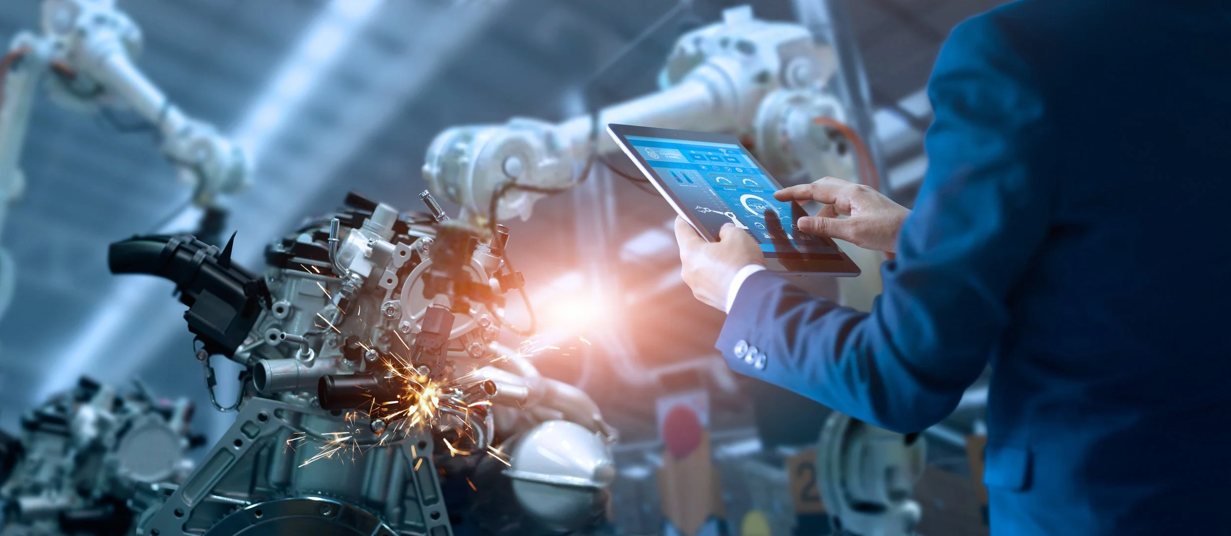 A person in a suit working on an engine with a digital tablet, sparks flying, in an industrial setting.