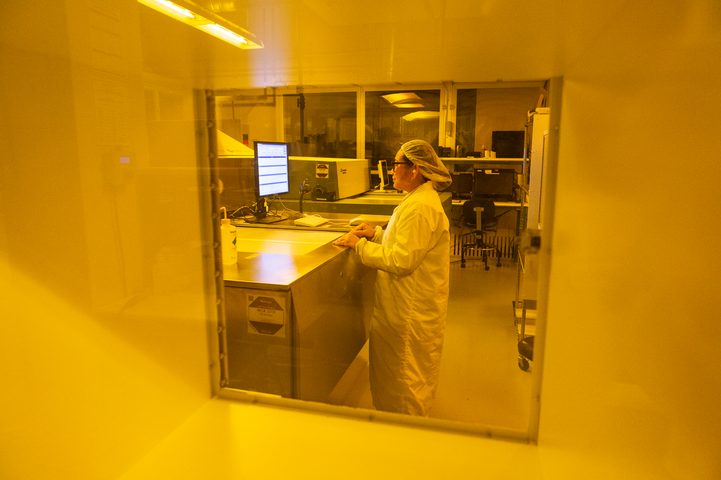 A person wearing a white lab coat, hairnet, and glasses working in a laboratory at a computer station, seen through a yellow-tinted window.