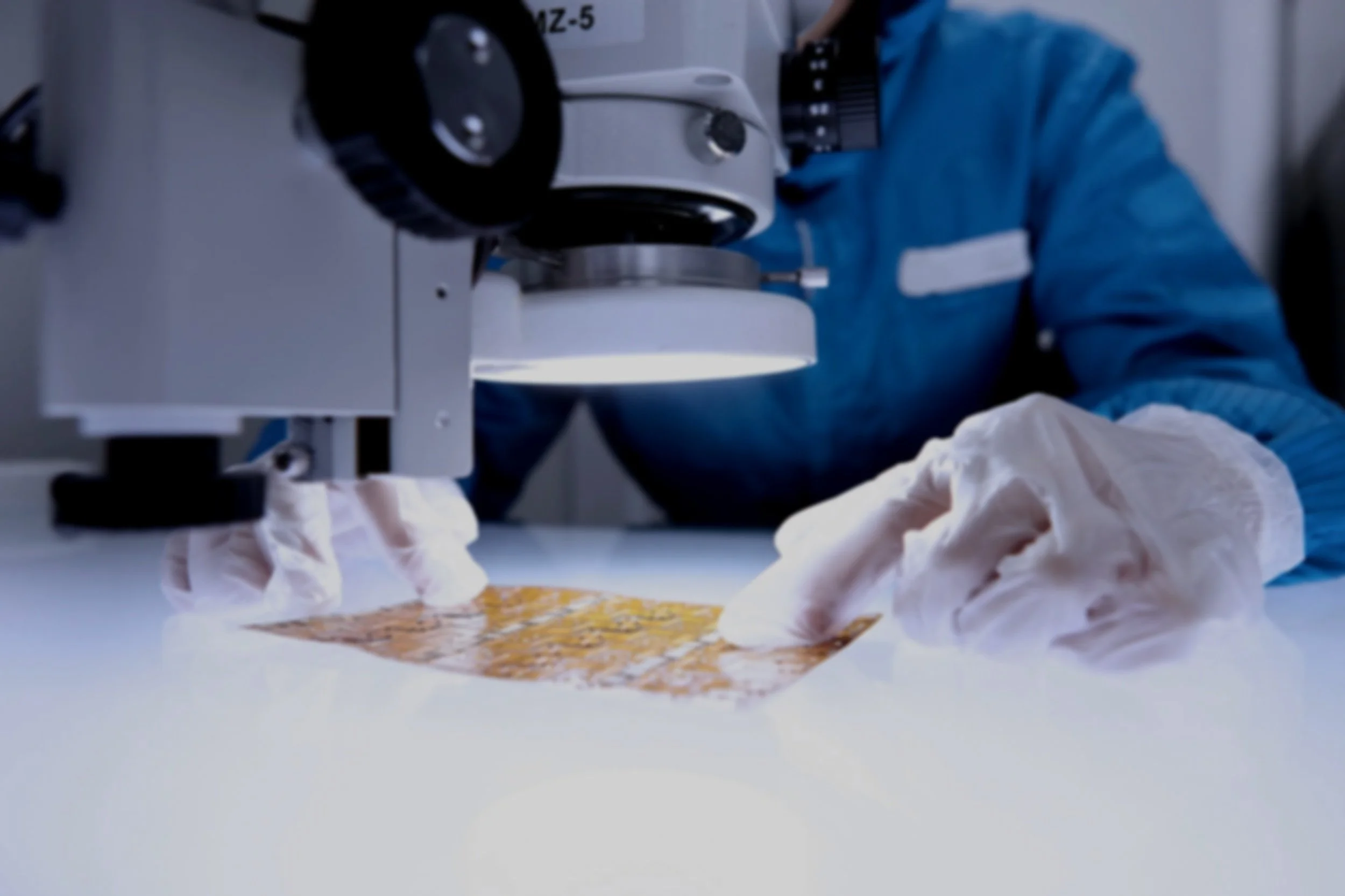 Close-up of a scientist in blue protective clothing using a microscope in a laboratory. The scientist's hands, covered with gloves, are working with a slide on the microscope's stage.