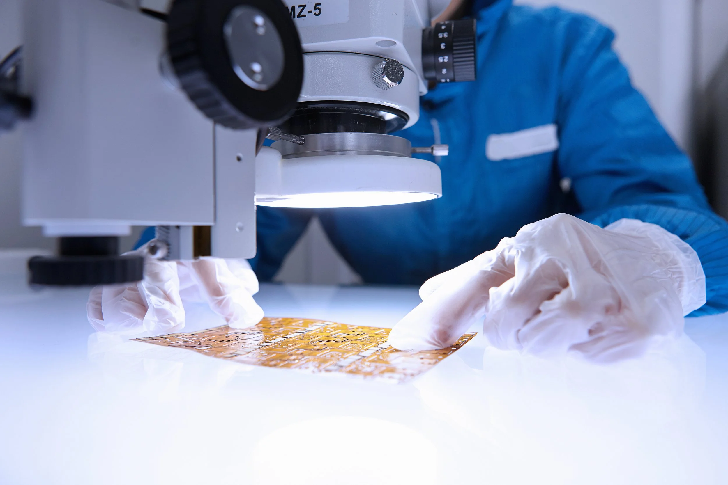 A scientist wearing blue protective clothing, gloves, and a lab coat is using a microscope to examine a sample placed on a slide.
