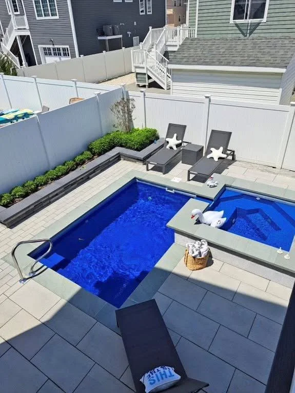 Backyard with a small pool and hot tub, two lounge chairs with star-shaped pillows, a small basket with towels, a swan-shaped pool float, and landscaped plants enclosed by white fencing.