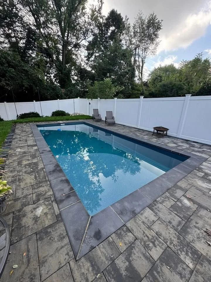 Backyard swimming pool with a white privacy fence, two lounge chairs, a small table, surrounded by gray stone pavers, with trees and a partly cloudy sky in the background.