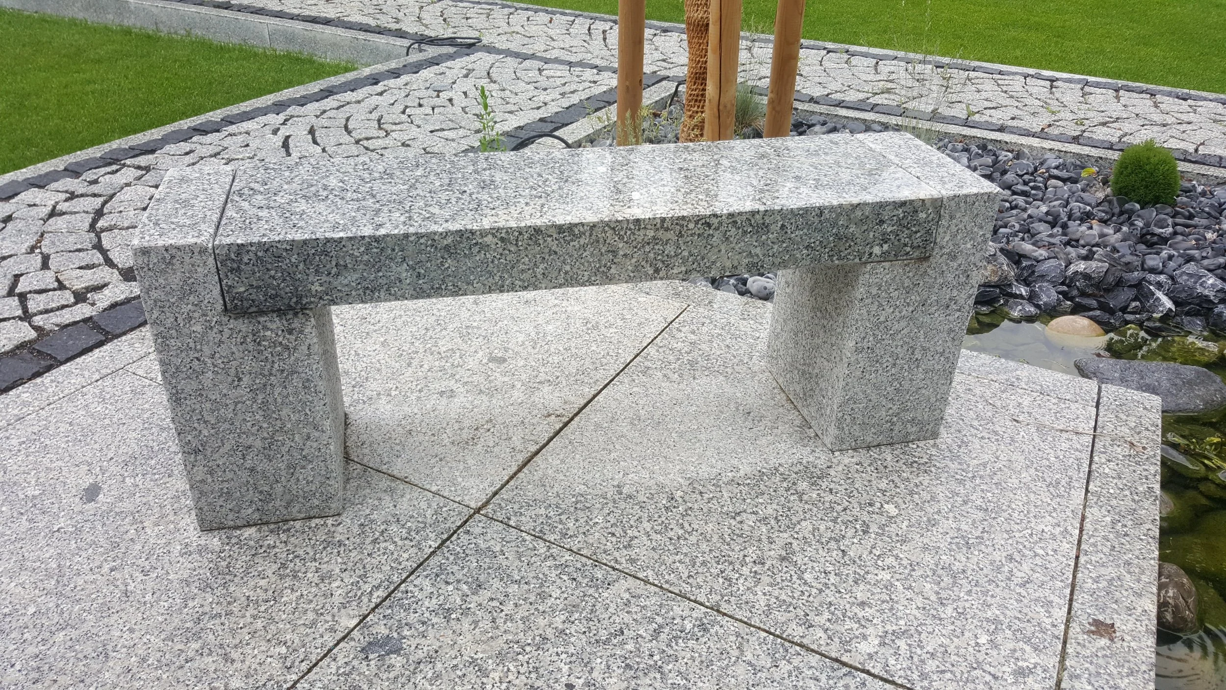 A granite bench situated on a paved outdoor surface near a small pond with rocks and plants.