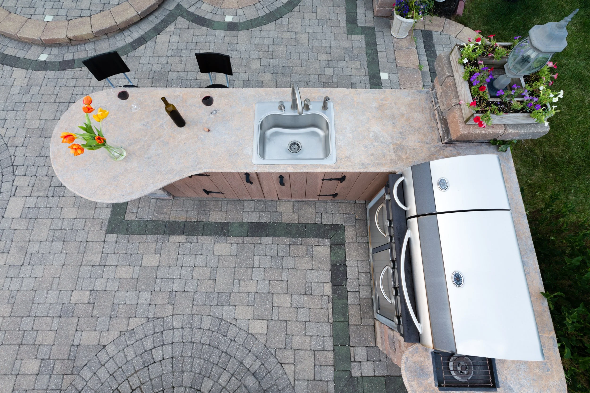 Outdoor kitchen area with a beige stone countertop, a sink, a small grill, colorful flowers in vases, a bottle of wine and two glasses of wine, surrounded by decorative brick and stone paving.