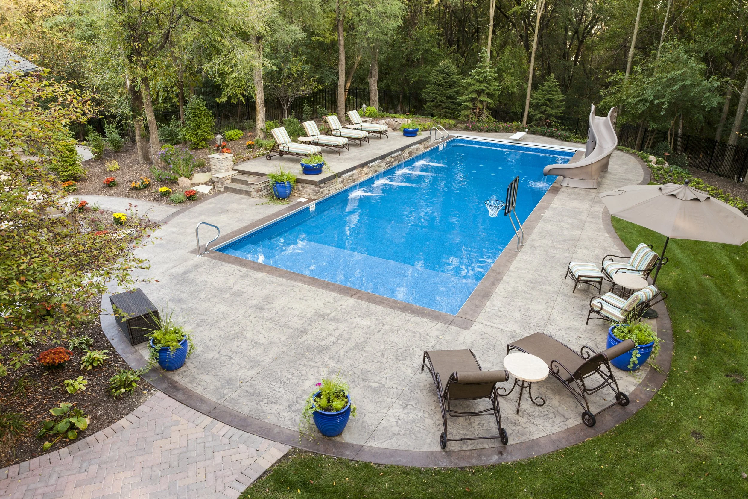 Residential backyard with a swimming pool, lounge chairs, a slide, potted plants, umbrellas, and surrounding trees and garden beds.