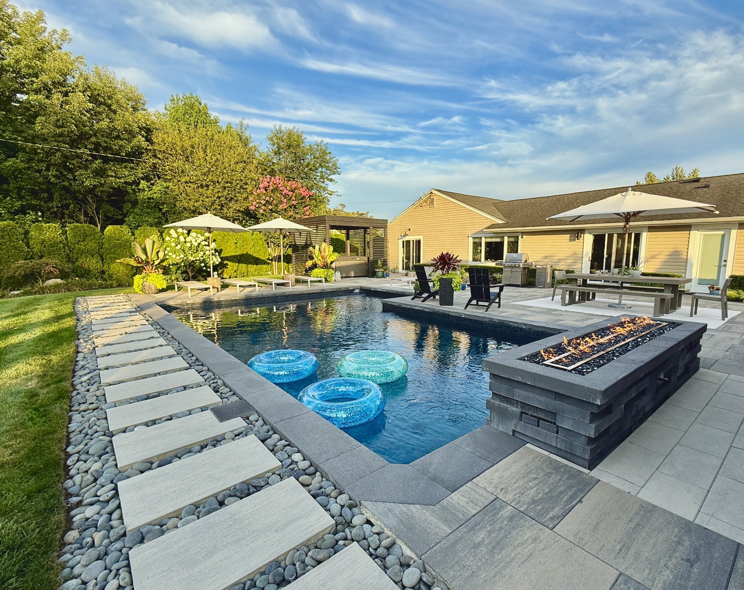 Backyard with swimming pool, poolside chairs, umbrellas, garden, grass, house with large windows, deck, trees, and blue sky with some clouds.