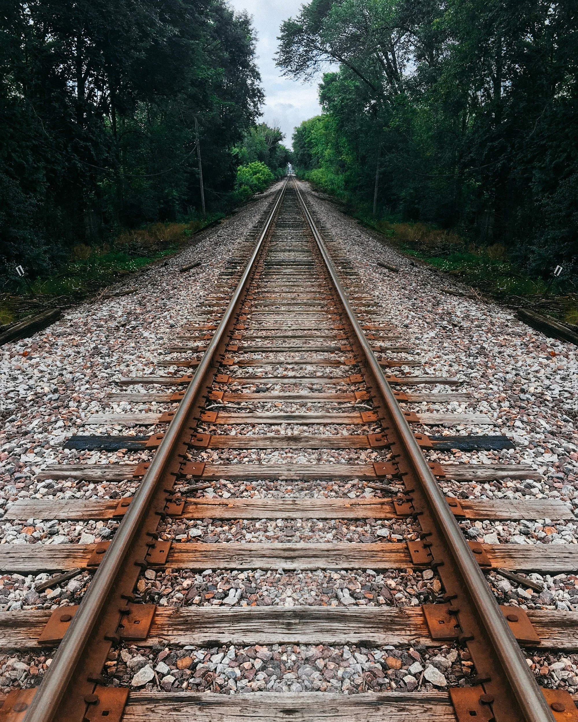 View of railroad tracks extending into the distance surrounded by lush green trees on both sides, under a cloudy sky.