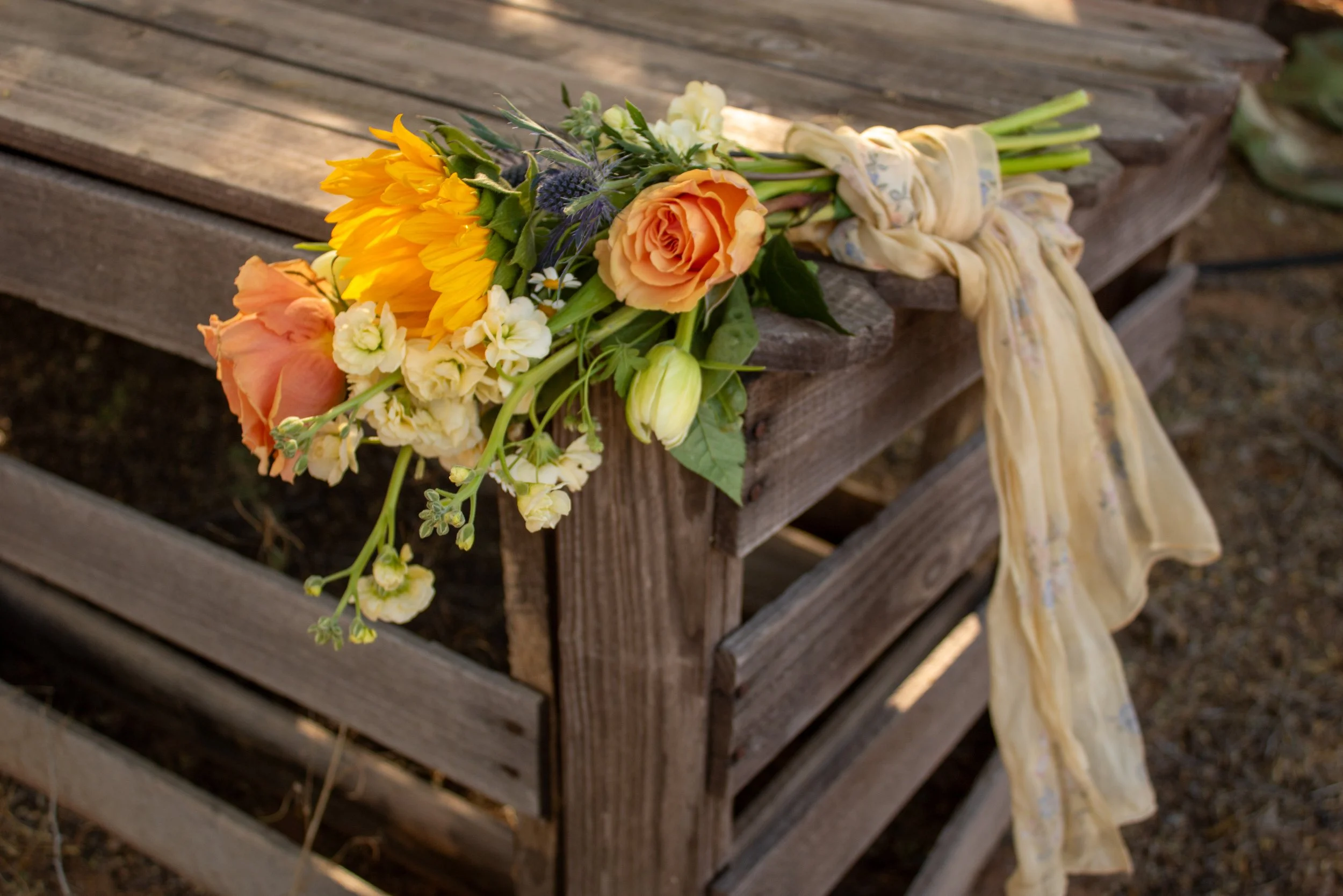 A bouquet of flowers, including orange roses and yellow sunflowers, tied with a light-colored fabric ribbon, rests on a weathered wooden bench outdoors.