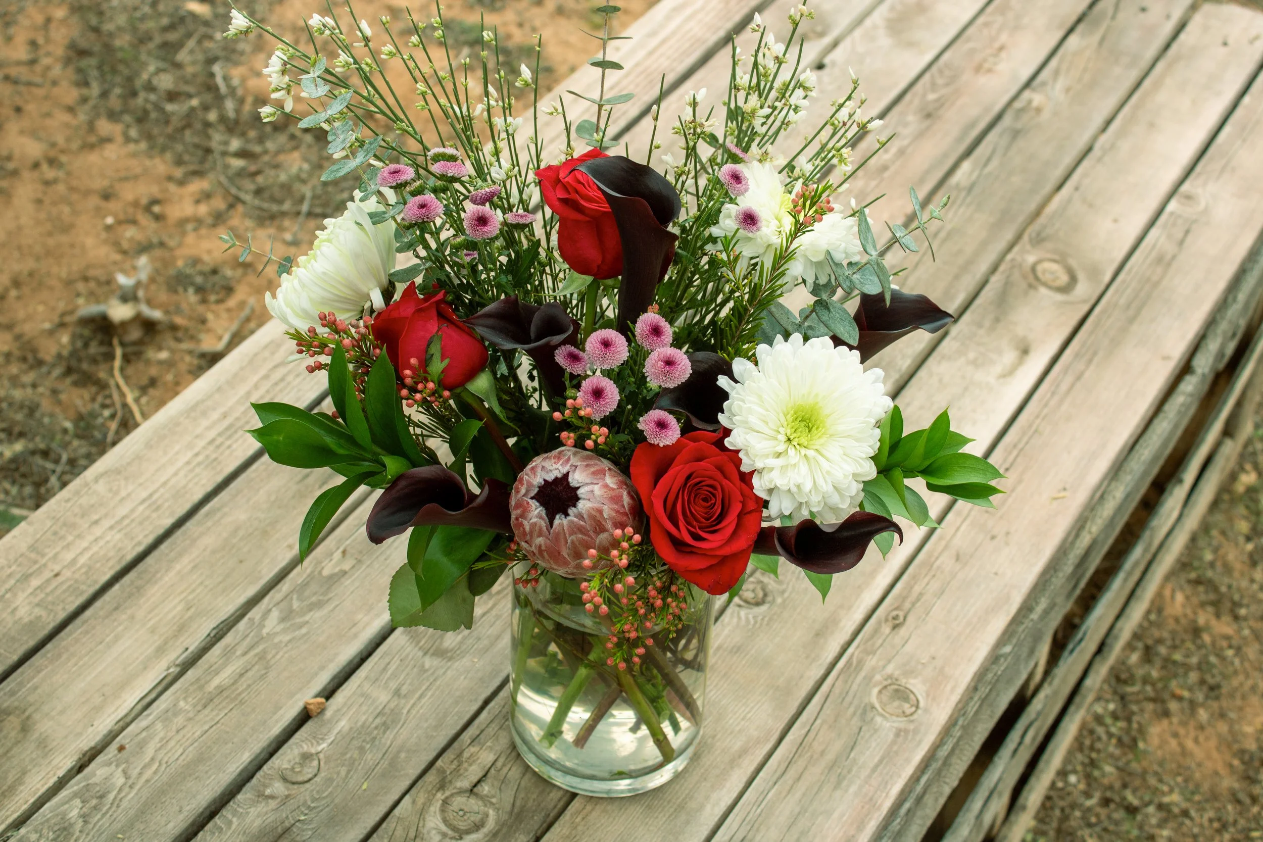 A glass vase filled with various flowers including red roses, white dahlias, dark calla lilies, pink button mums, and greenery, placed on a wooden surface outdoors.