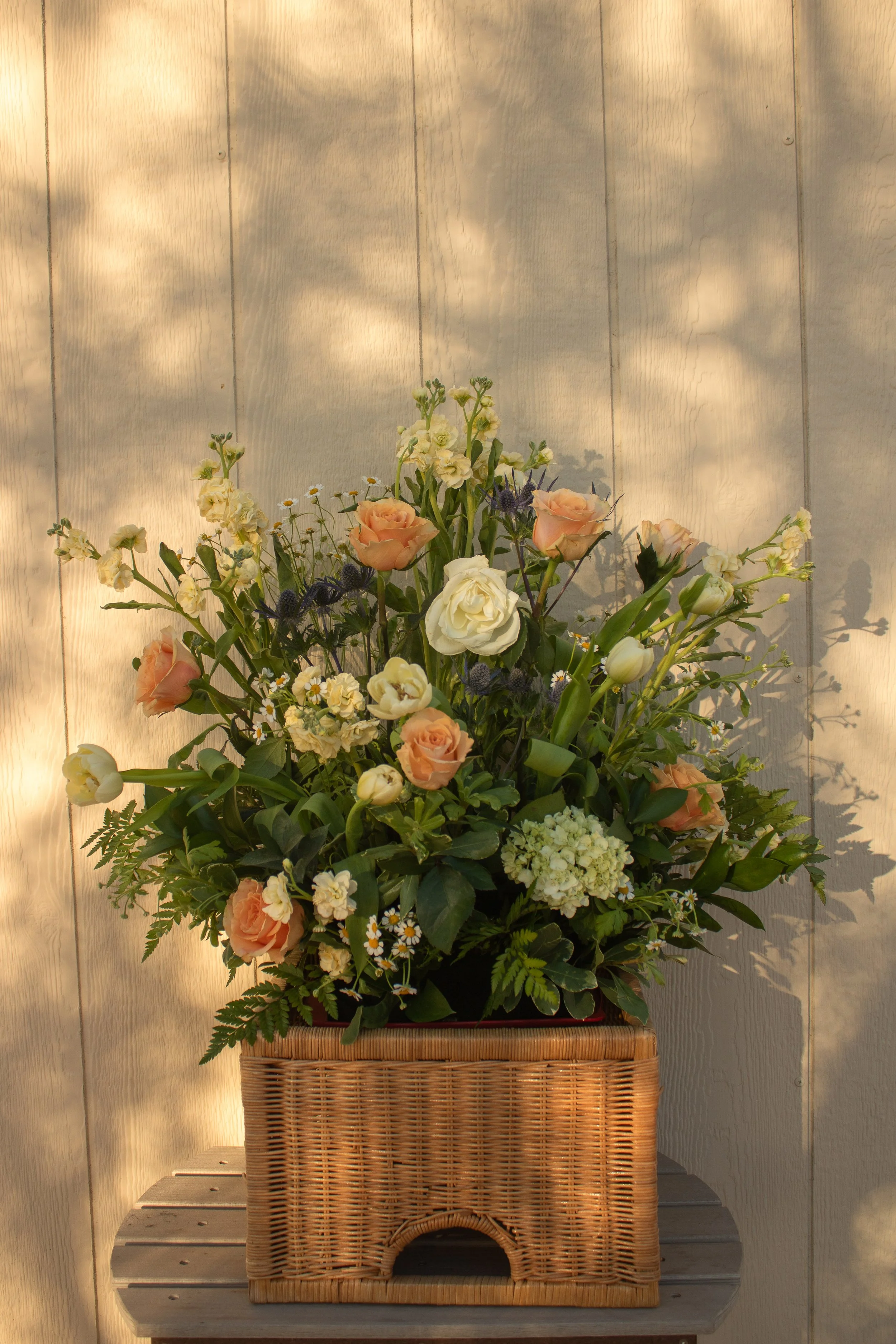 A floral arrangement with light peach, cream, and white flowers, as well as small daisies, placed in a basket with a woven design, against a light wooden wall with shadows.