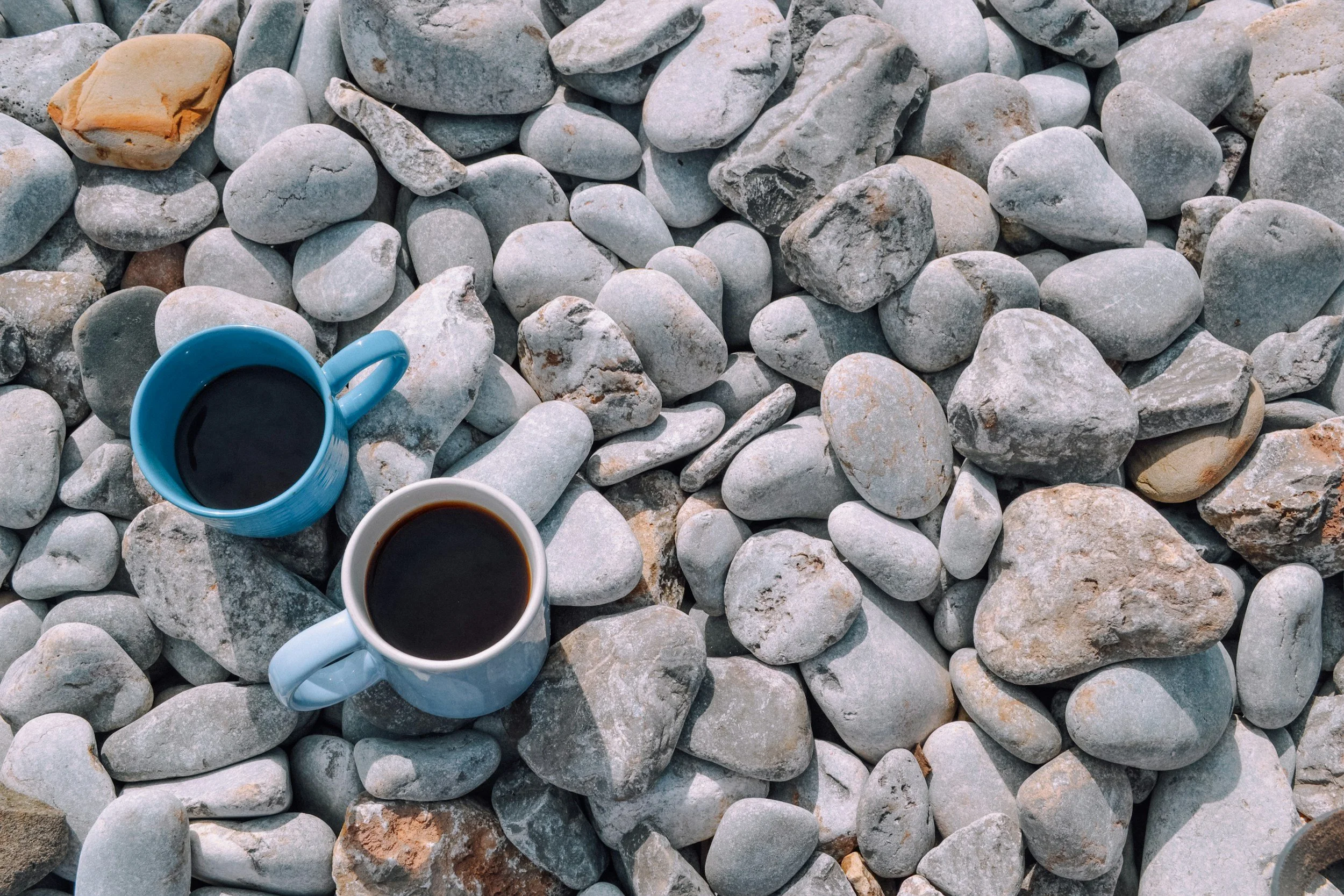Two cups of black coffee on a bed of gray and tan stones, with a light blue mug in the foreground and a white mug partially visible in the bottom right corner.