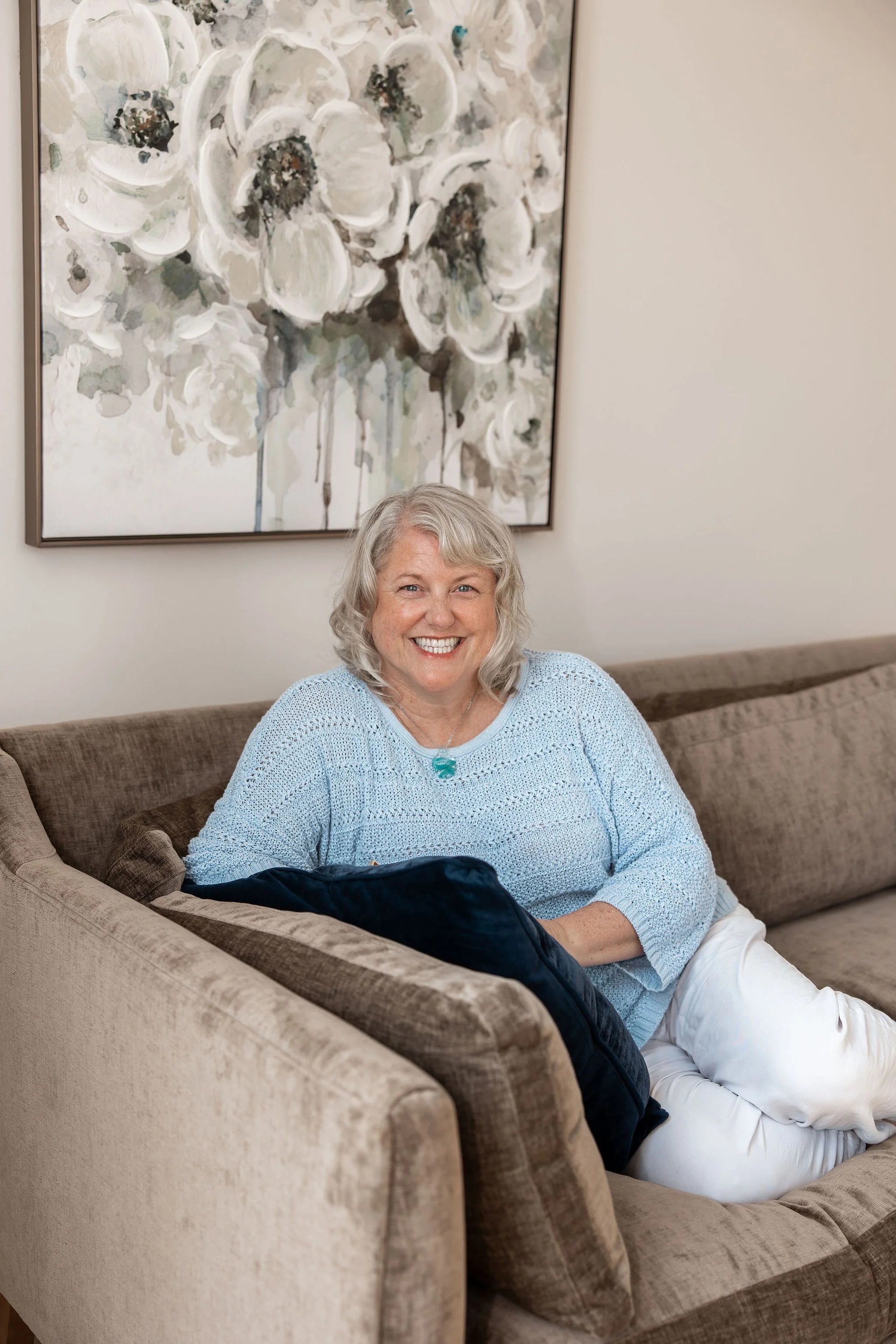 A smiling woman with gray hair sitting on a beige couch with a dark cushion, in a room with a large abstract floral painting on the wall behind her.