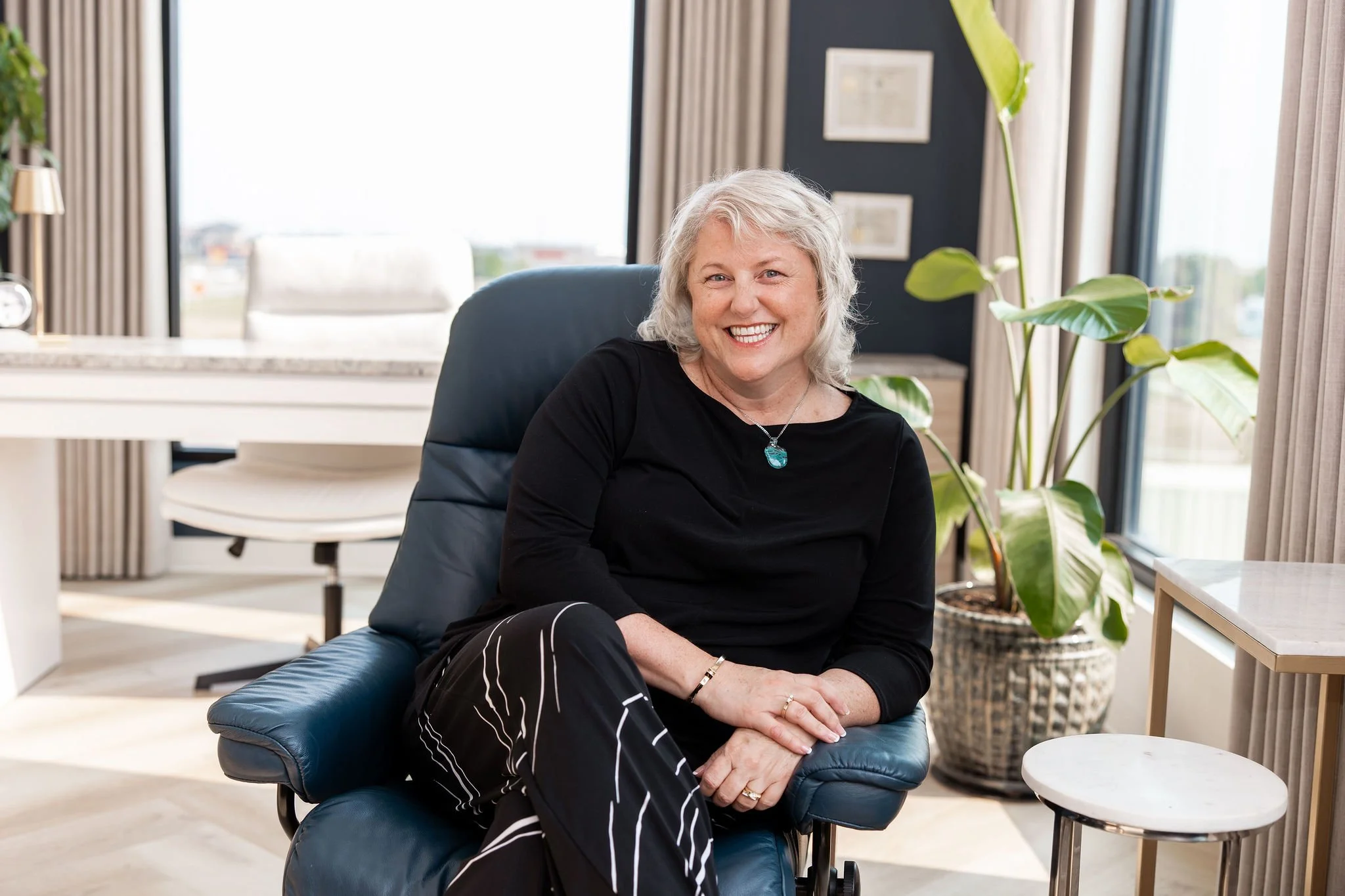 A smiling woman with gray hair sitting in a black armchair in a modern, well-lit office space with large windows, a potted plant, and framed artwork on the wall.