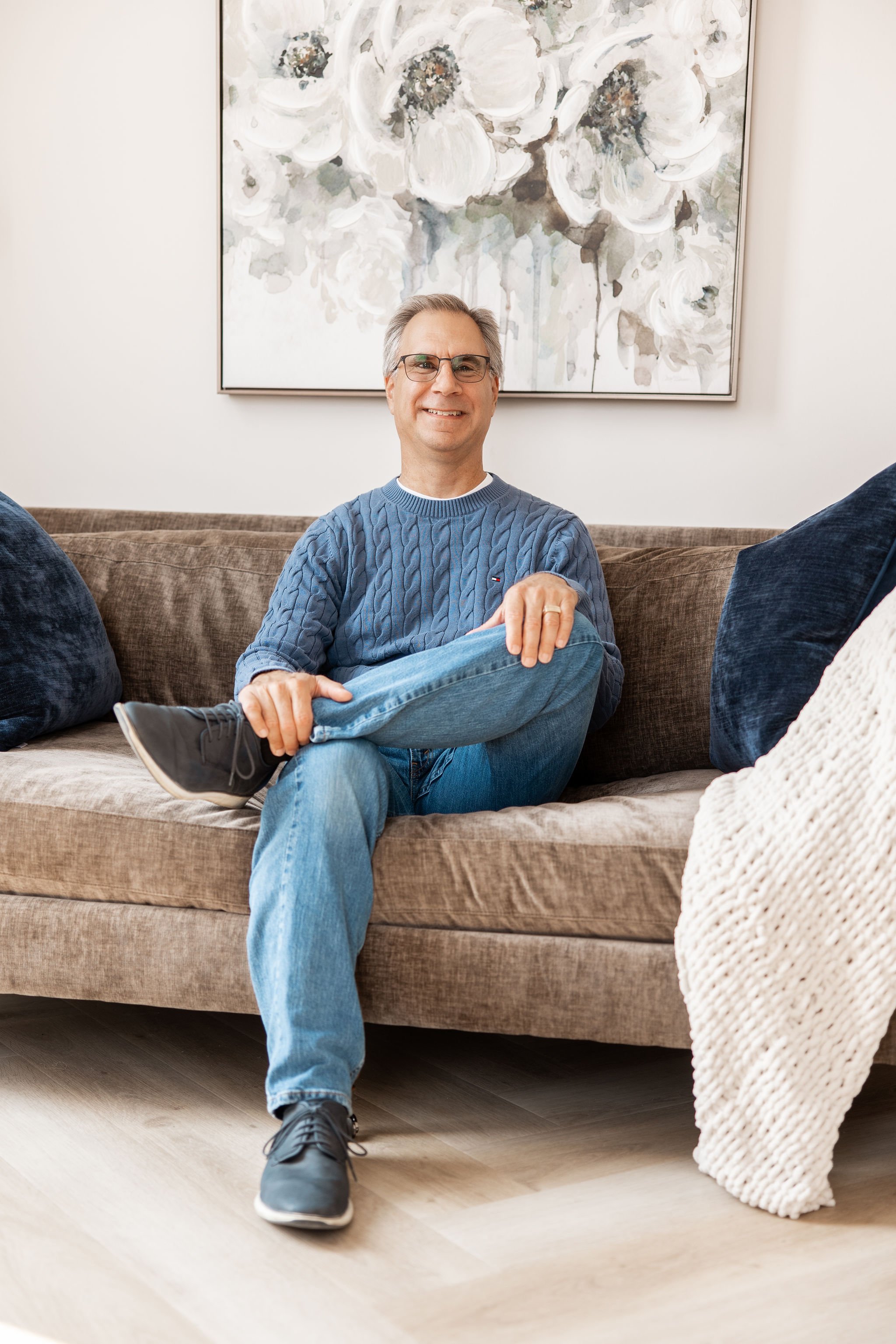 A smiling man with glasses and gray hair, wearing a blue cable-knit sweater, sitting on a brown sofa in front of a white wall with abstract floral artwork, with his left leg crossed over his right knee and his left hand resting on his ankle.