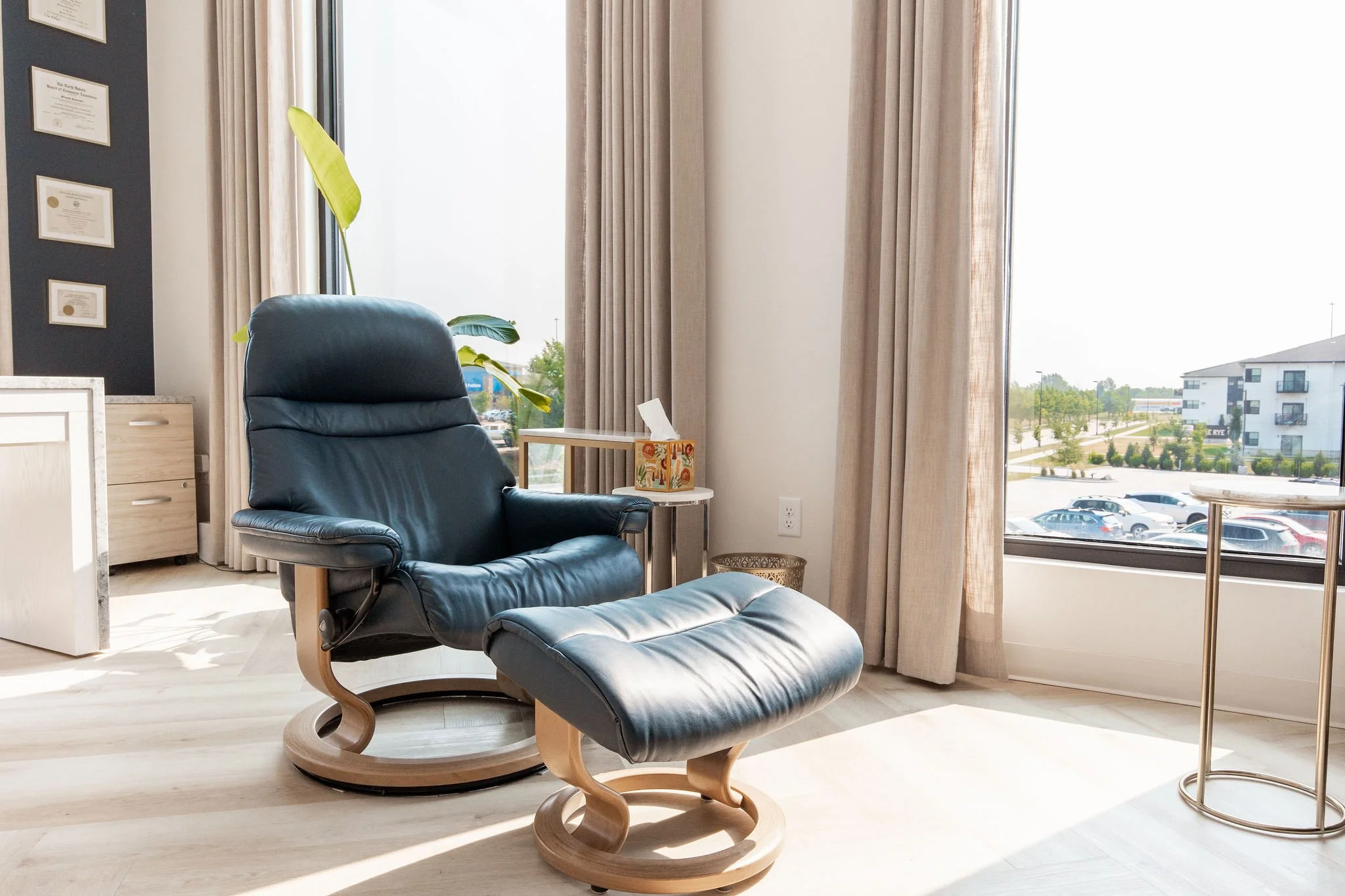 Modern office or living space with a black leather lounge chair and matching ottoman, natural light streaming through large windows with beige curtains, and framed certificates on a dark wall.