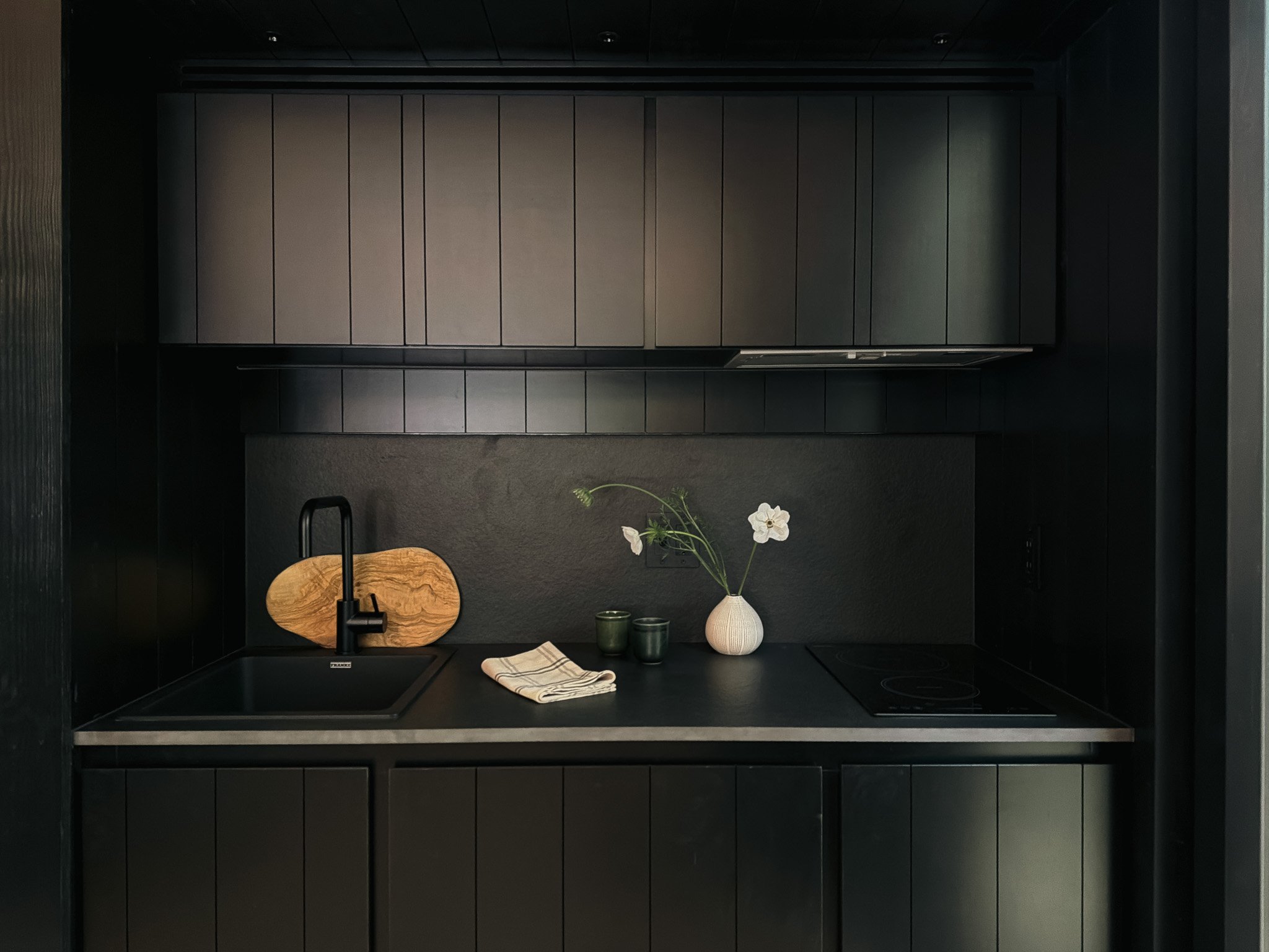 A modern kitchen with dark cabinetry, a black countertop, and a black backsplash. There is a white vase with flowers, two small green containers, a folded cloth, and a wooden cutting board on the counter.