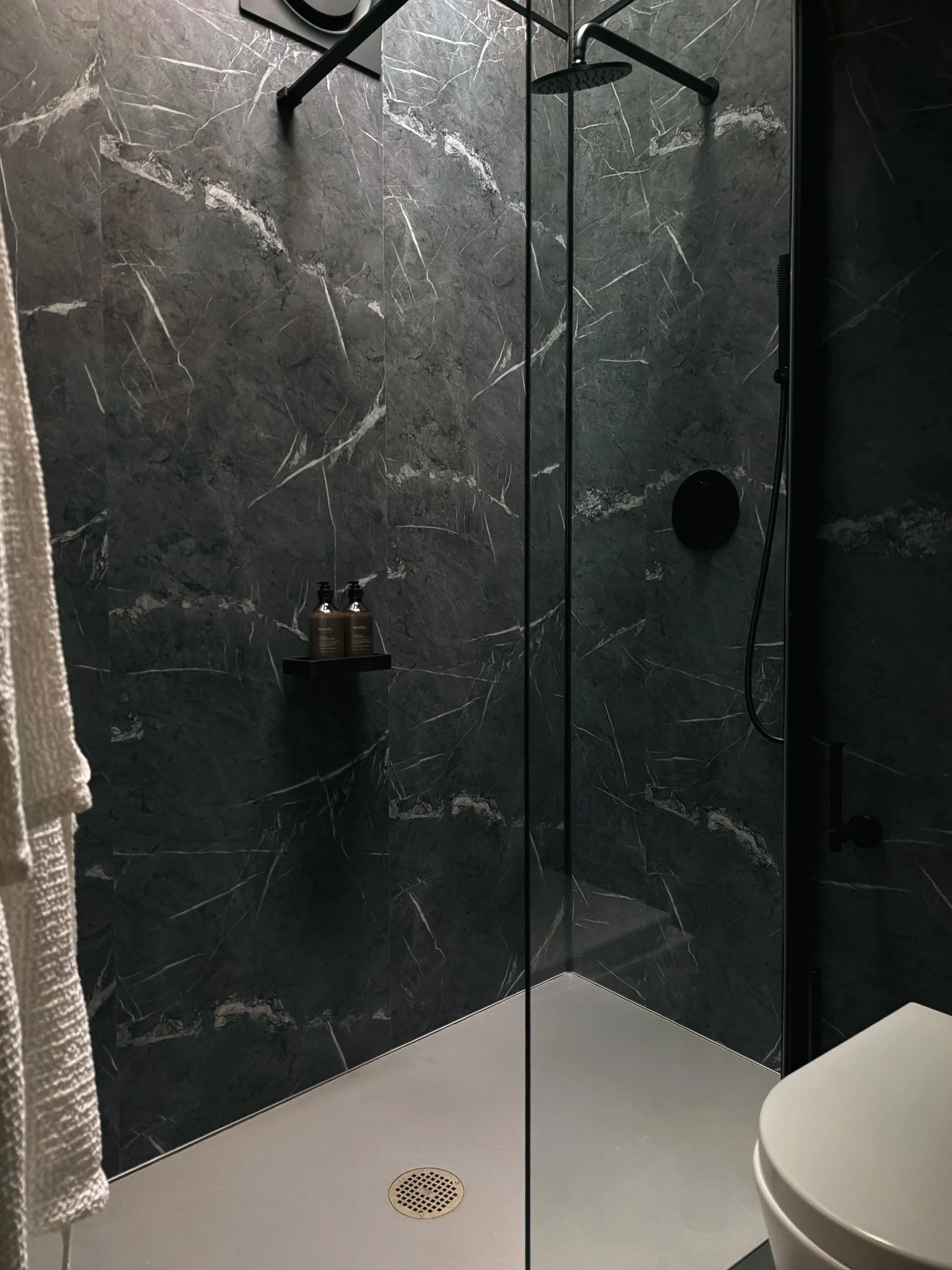 A modern bathroom with black marble walls, a walk-in shower with a black showerhead, and a small shelf holding shampoo bottles. A white toilet is visible in the foreground, and a towel hangs on a hook to the left.