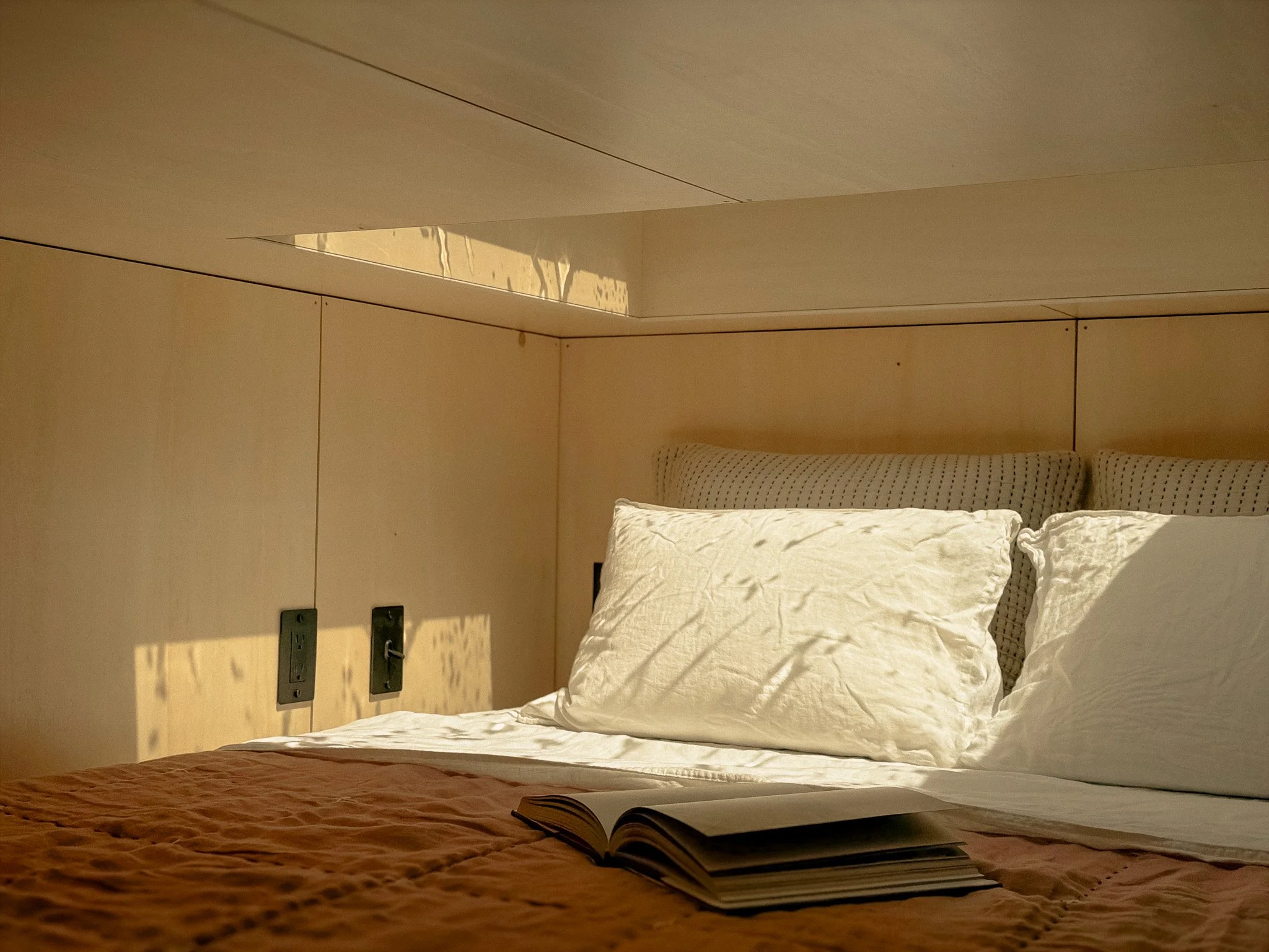 A bedroom with beige walls and bedding. The bed has white pillows and a book open on a brown quilted coverlet. There is a headboard with built-in lighting and electrical outlets.