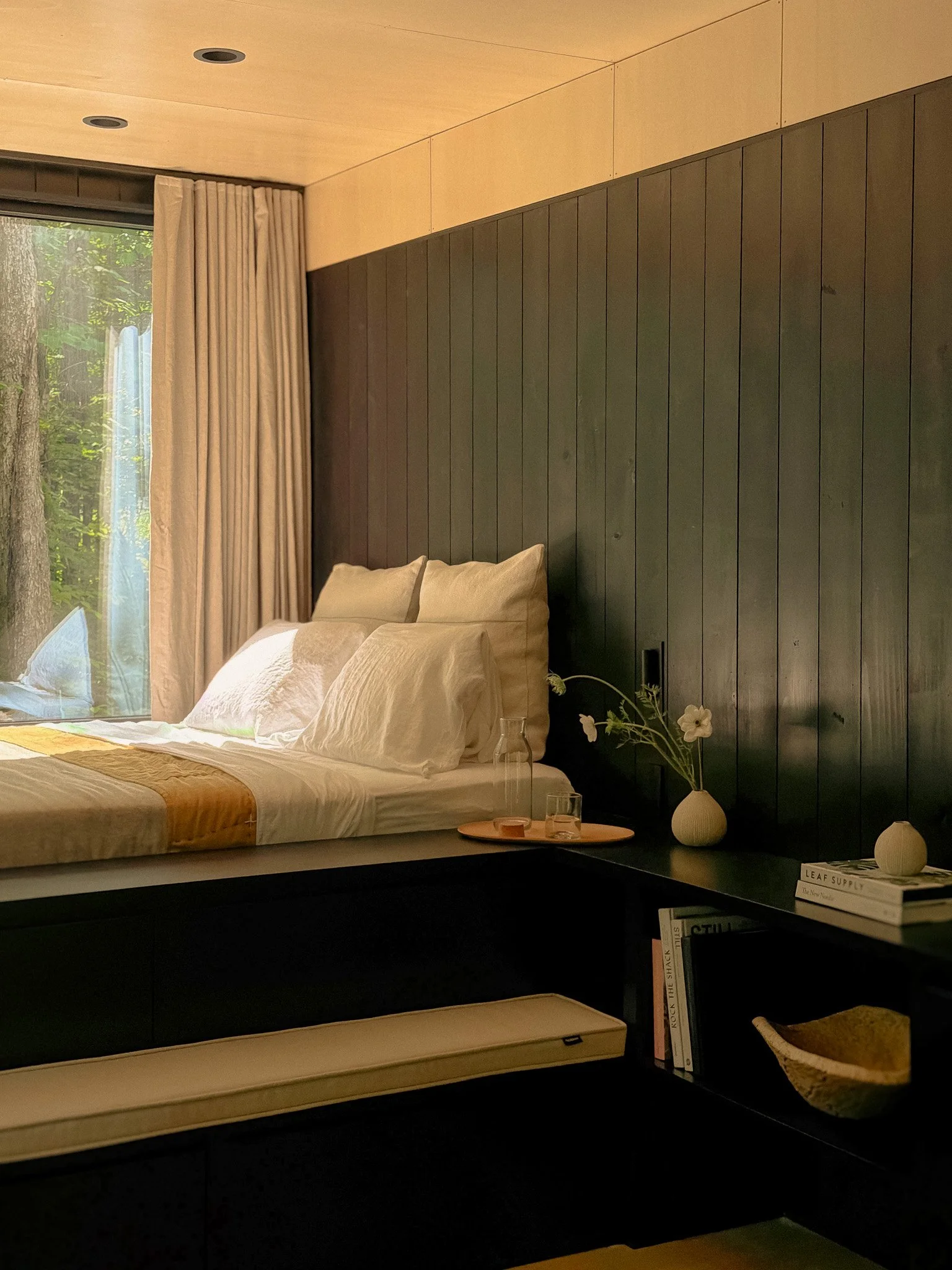 A bedroom with a bed that has white pillows and a beige quilt, against a black wooden paneled wall. A large window with beige curtains shows trees outside. On the black shelf below the bed, there are a few books, a small white vase with flowers, and a tray with glasses and a candle.