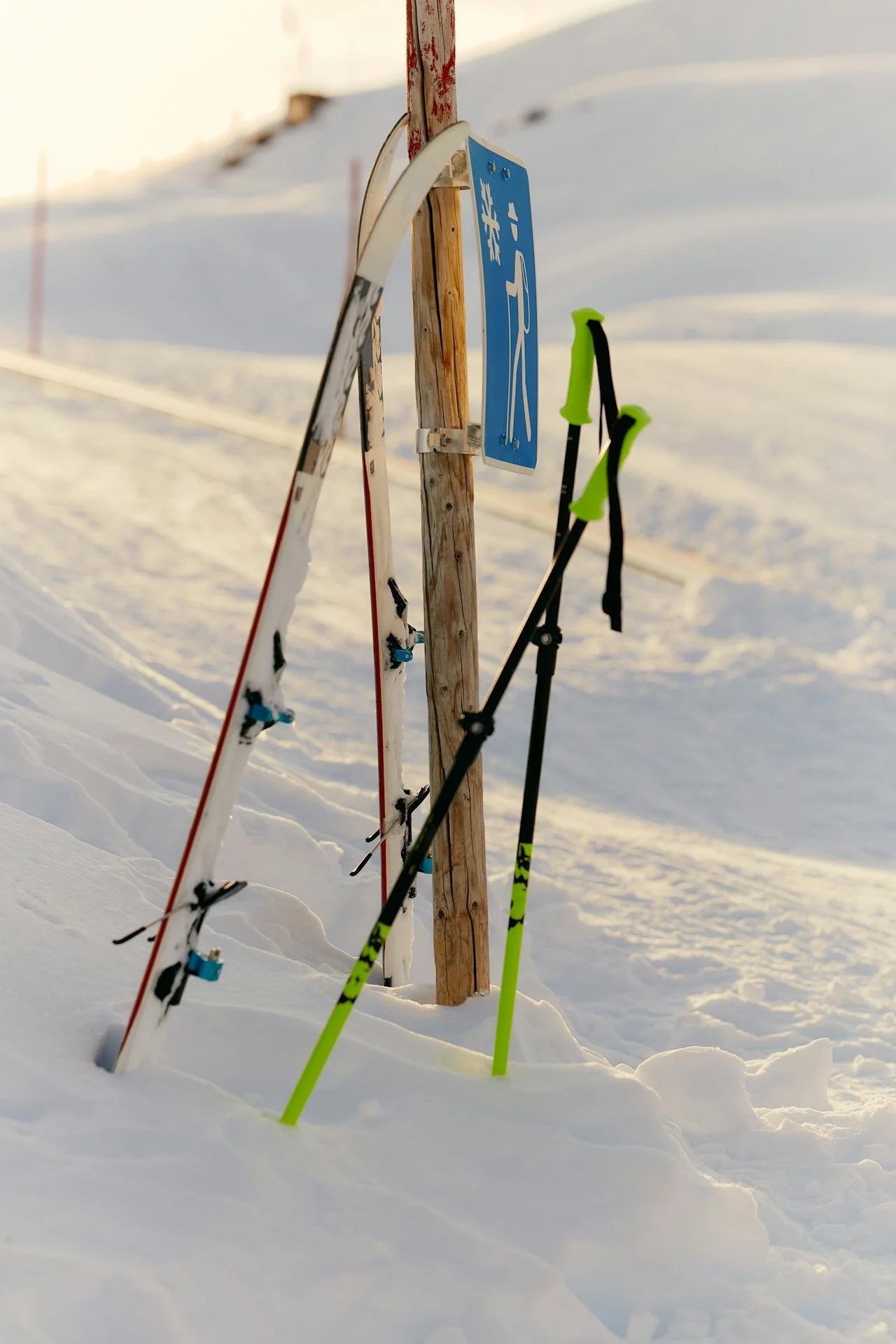 Ski poles and skis propped against a wooden post in the snow with a blue ski trail sign in the background at a snowy outdoor location, likely a ski resort or trail.