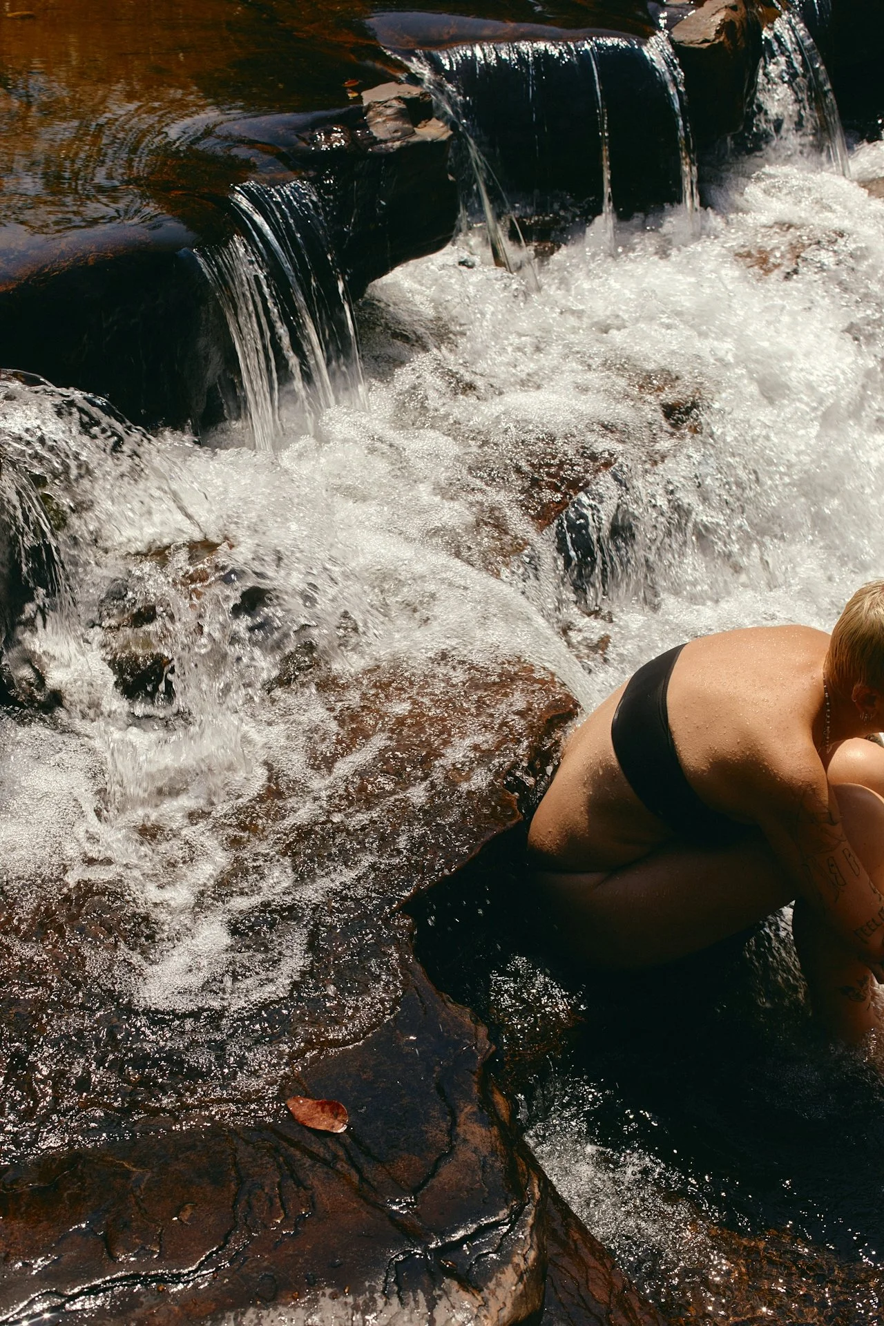 A person in a black swimsuit sitting on rocks in a river with rushing water, with water cascading over the rocks, and part of the person's arm and side visible.