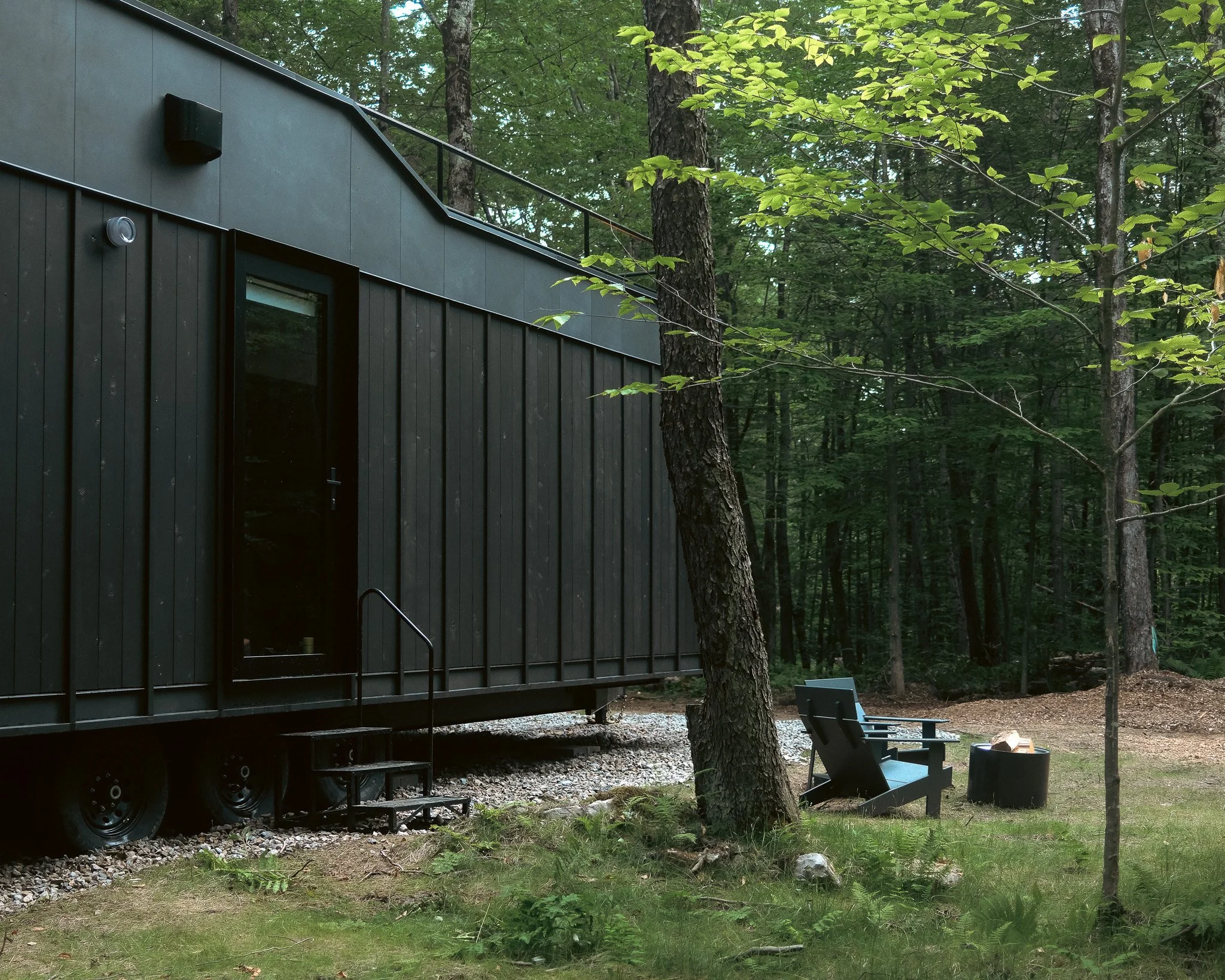 A modern black cabin in a forest with tall trees and green leaves. The cabin has a large window and a small staircase leading to the door. There are chairs and a fire pit in front of the cabin.