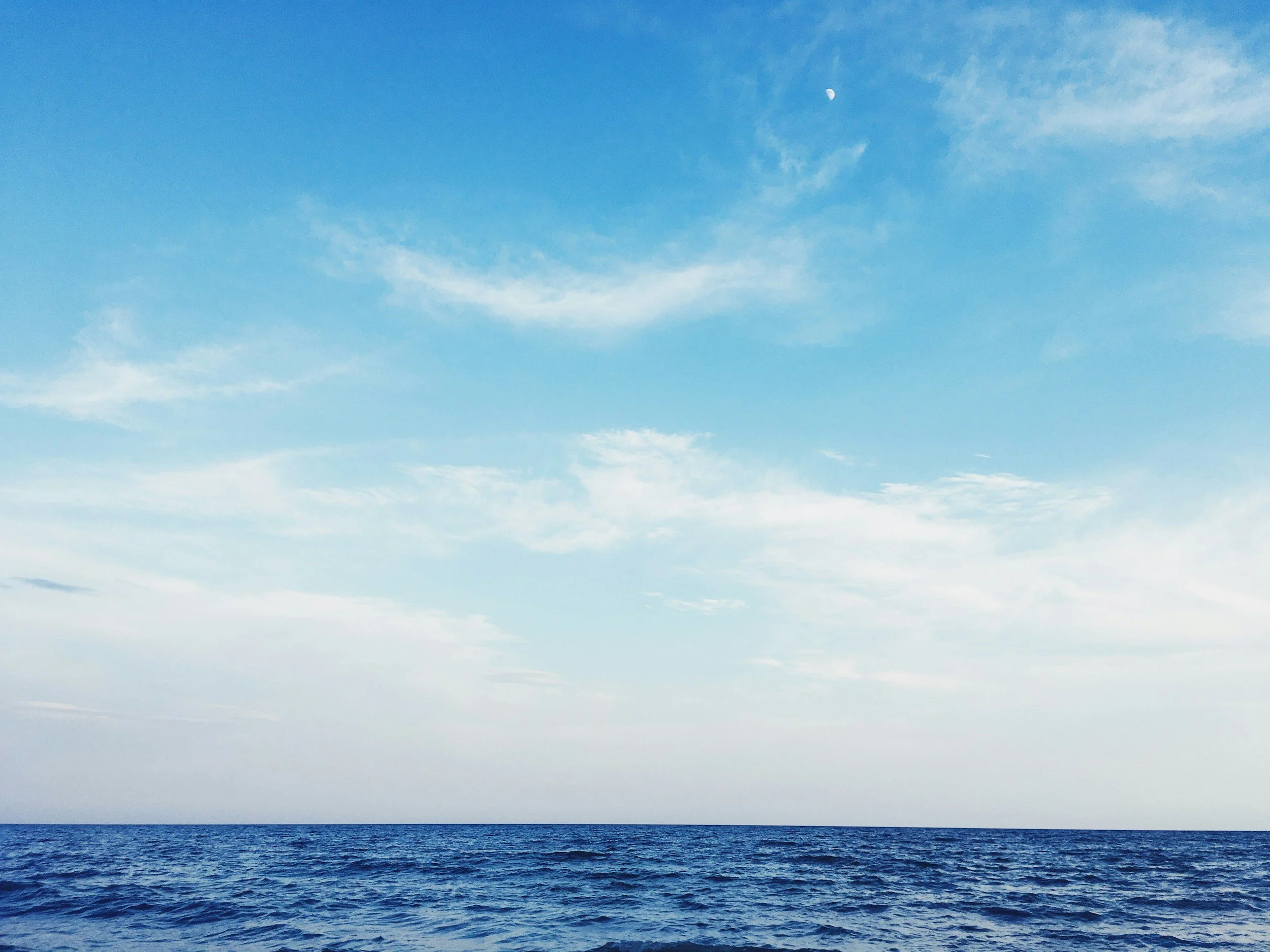 View of the ocean with a clear blue sky and some clouds, and the moon visible in the sky.