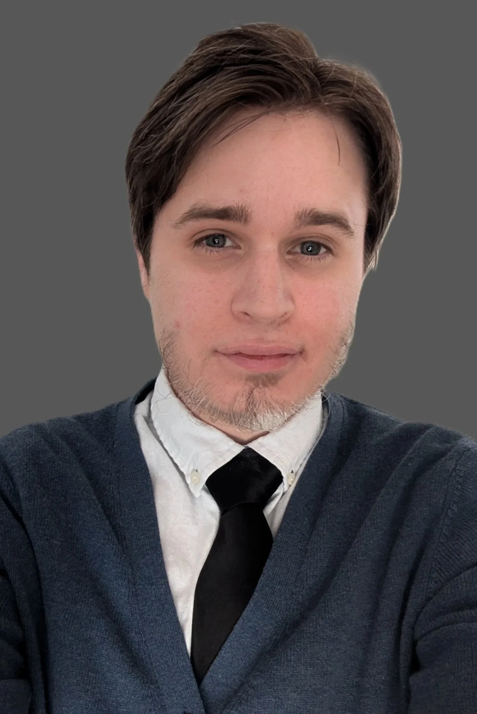A young man with short brown hair, light beard, and blue eyes, wearing a white shirt, black tie, and a dark blue sweater, taking a selfie against a gray background.