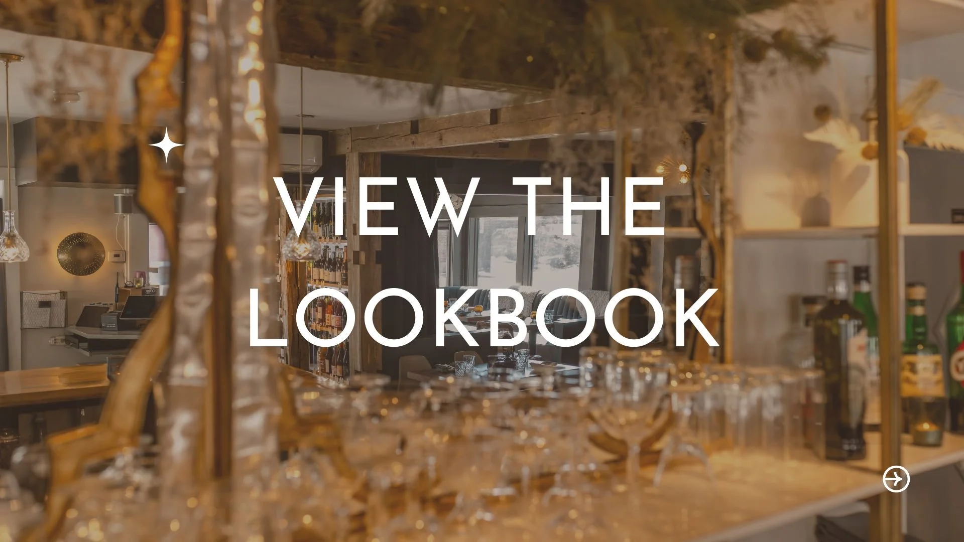 Interior of a restaurant or bar with wine bottles, glasses, and modern decor, viewed through a mirror or glass partition with text overlay that reads "VIEW THE LOOKBOOK."