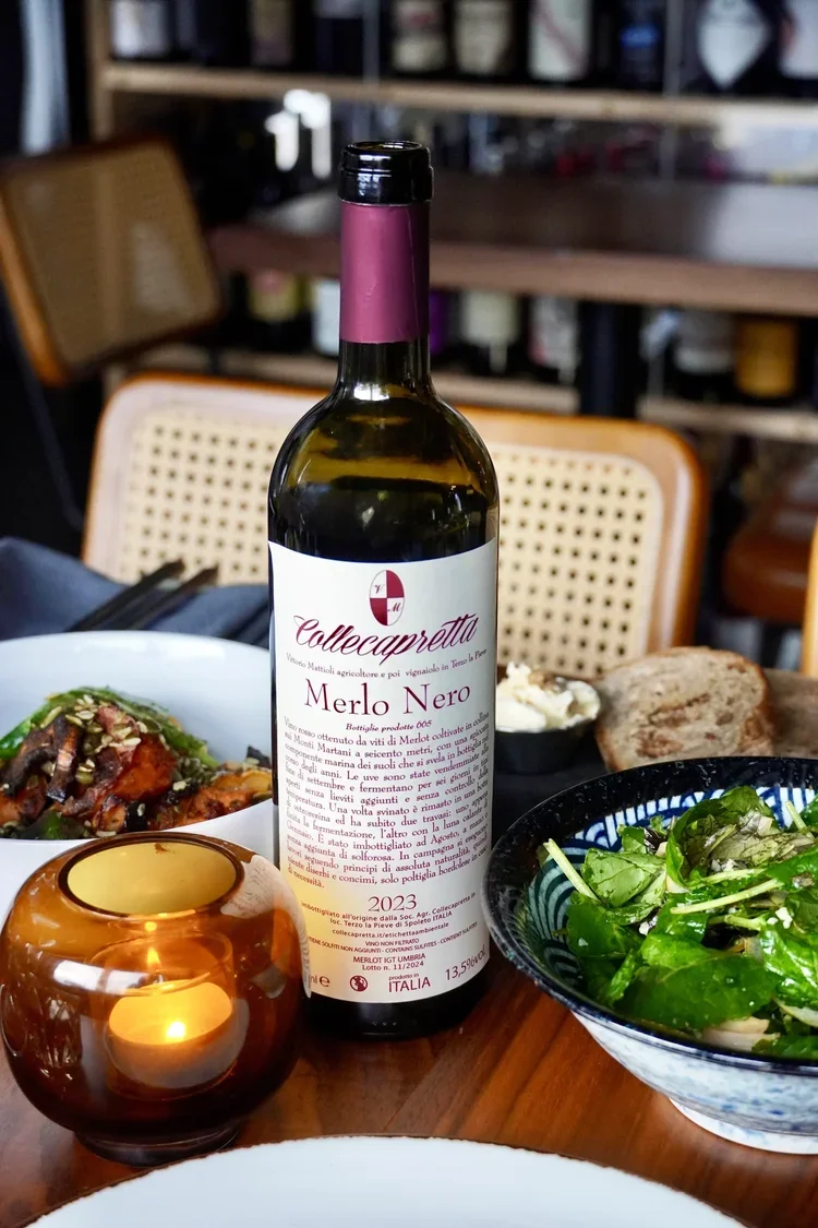 A bottle of Collecaprette Merlo Nero red wine on a dinner table, surrounded by a salad, a candle in a glass holder, bread, and a bowl with some spread in a cozy restaurant setting at Cork Restaurant