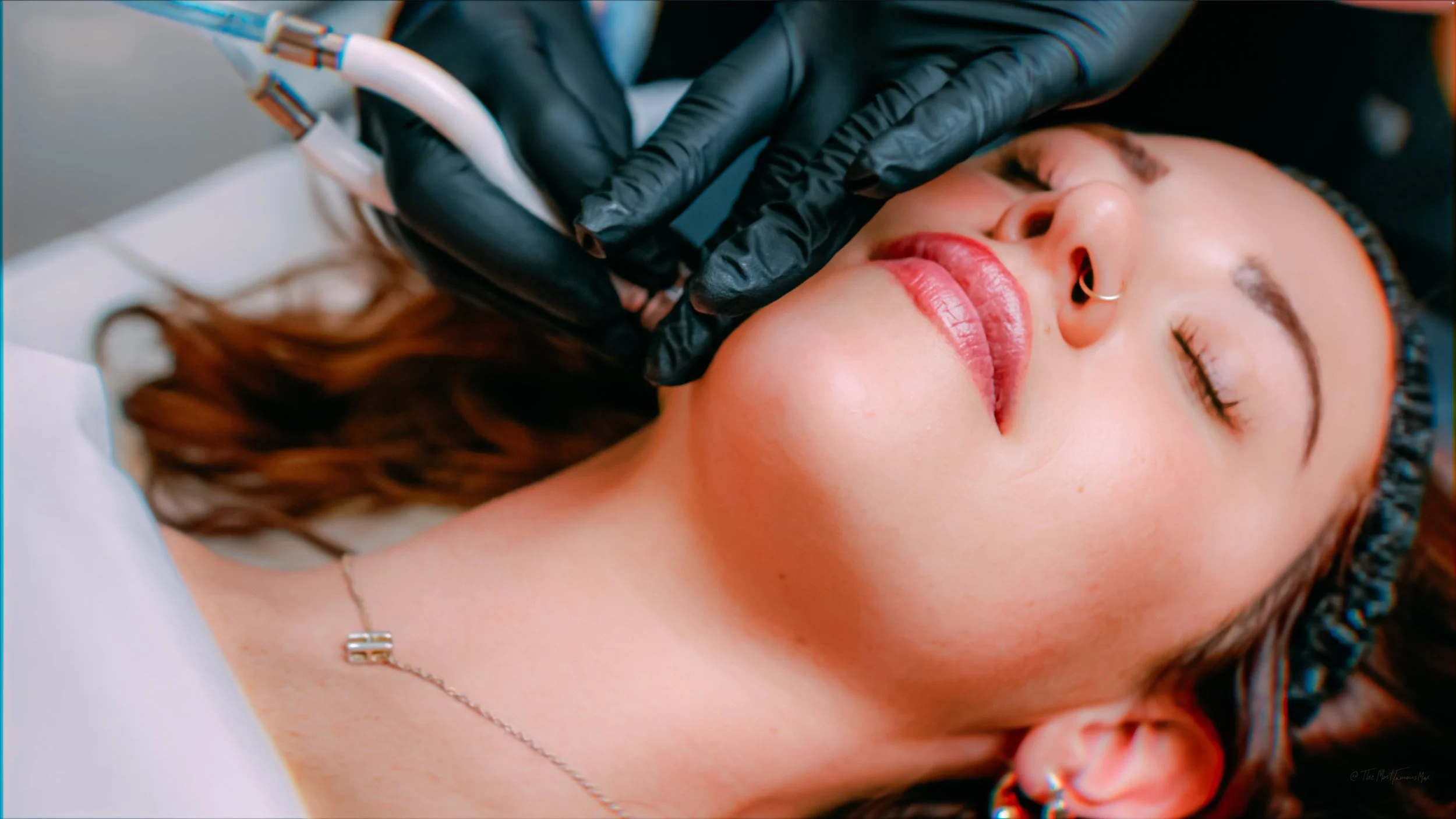 A woman with closed eyes receiving a facial treatment from a person wearing black gloves holding a device near her face.