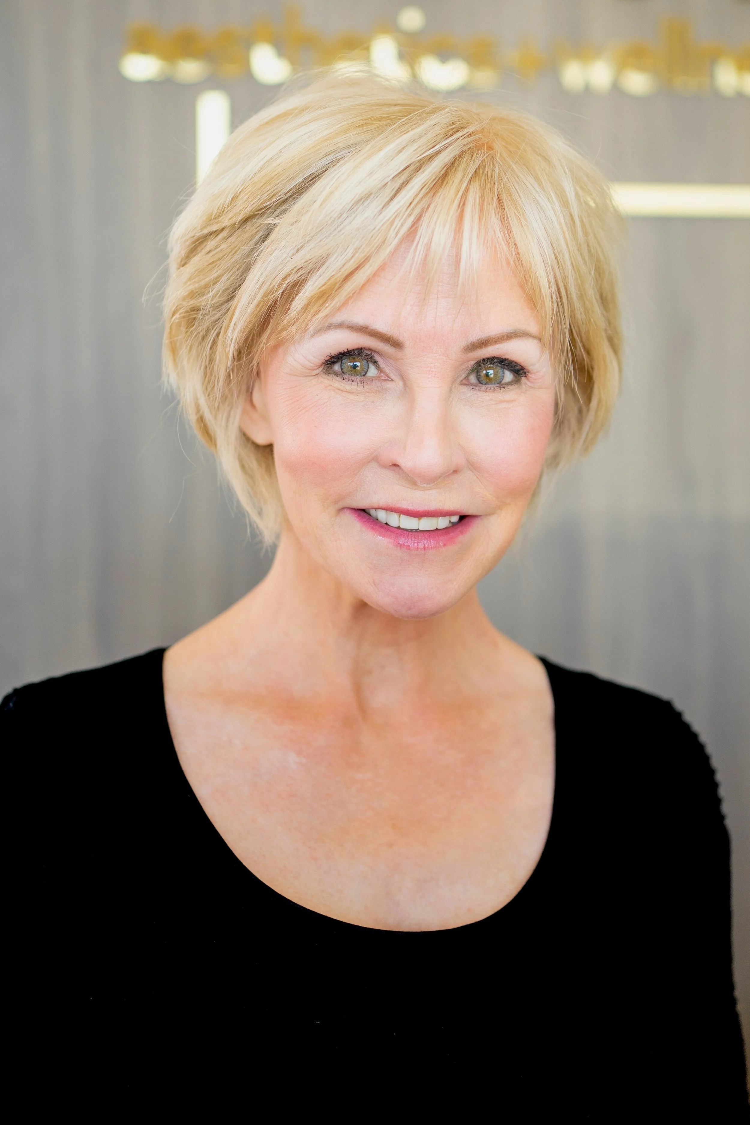 Close-up photograph of a middle-aged woman with short blonde hair, wearing a black top, smiling, against a blurred gray background.