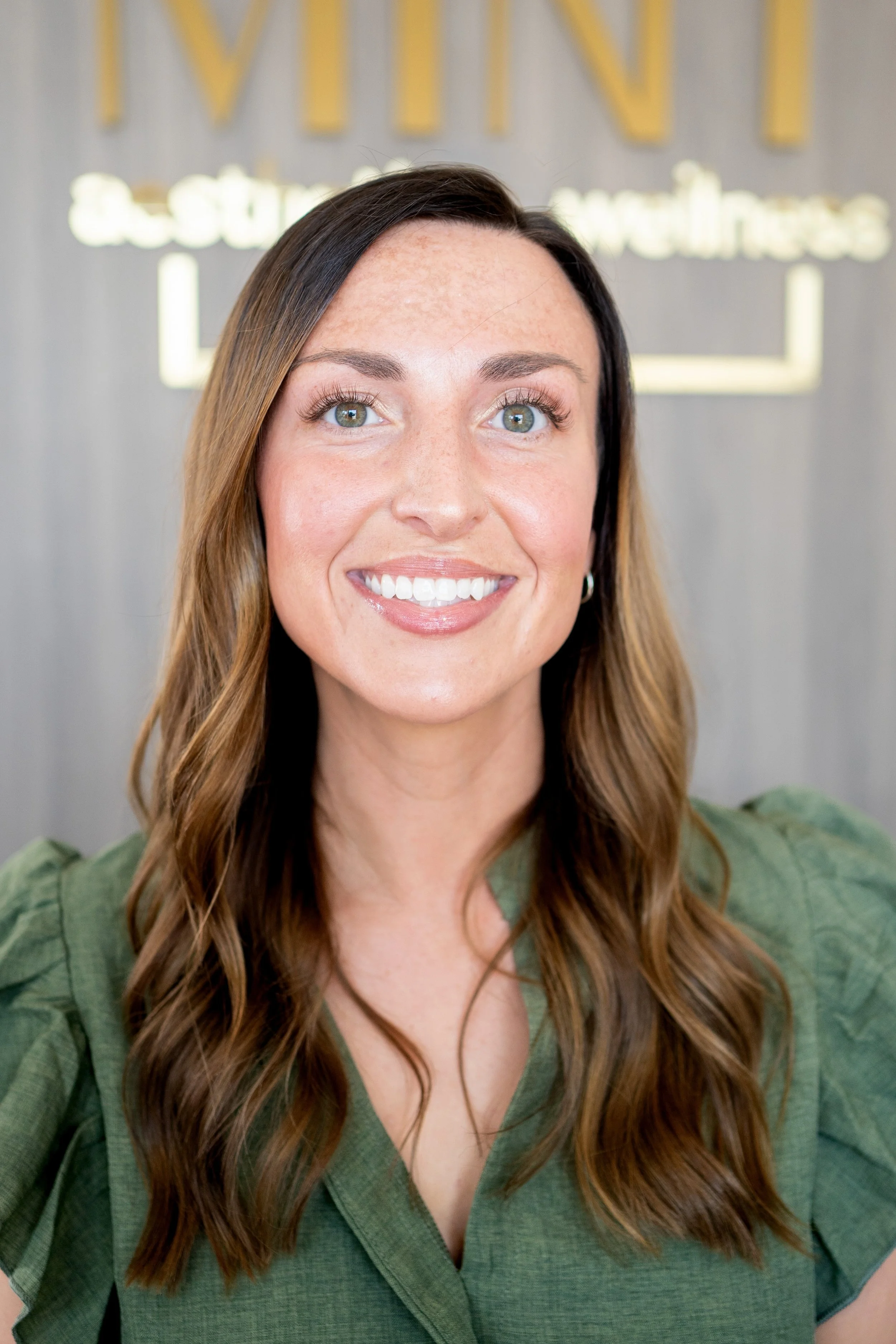 A woman with long brown hair smiling, wearing a green shirt, standing in front of a wall with partial sign including the words 'VINT' and 'esthetics'.