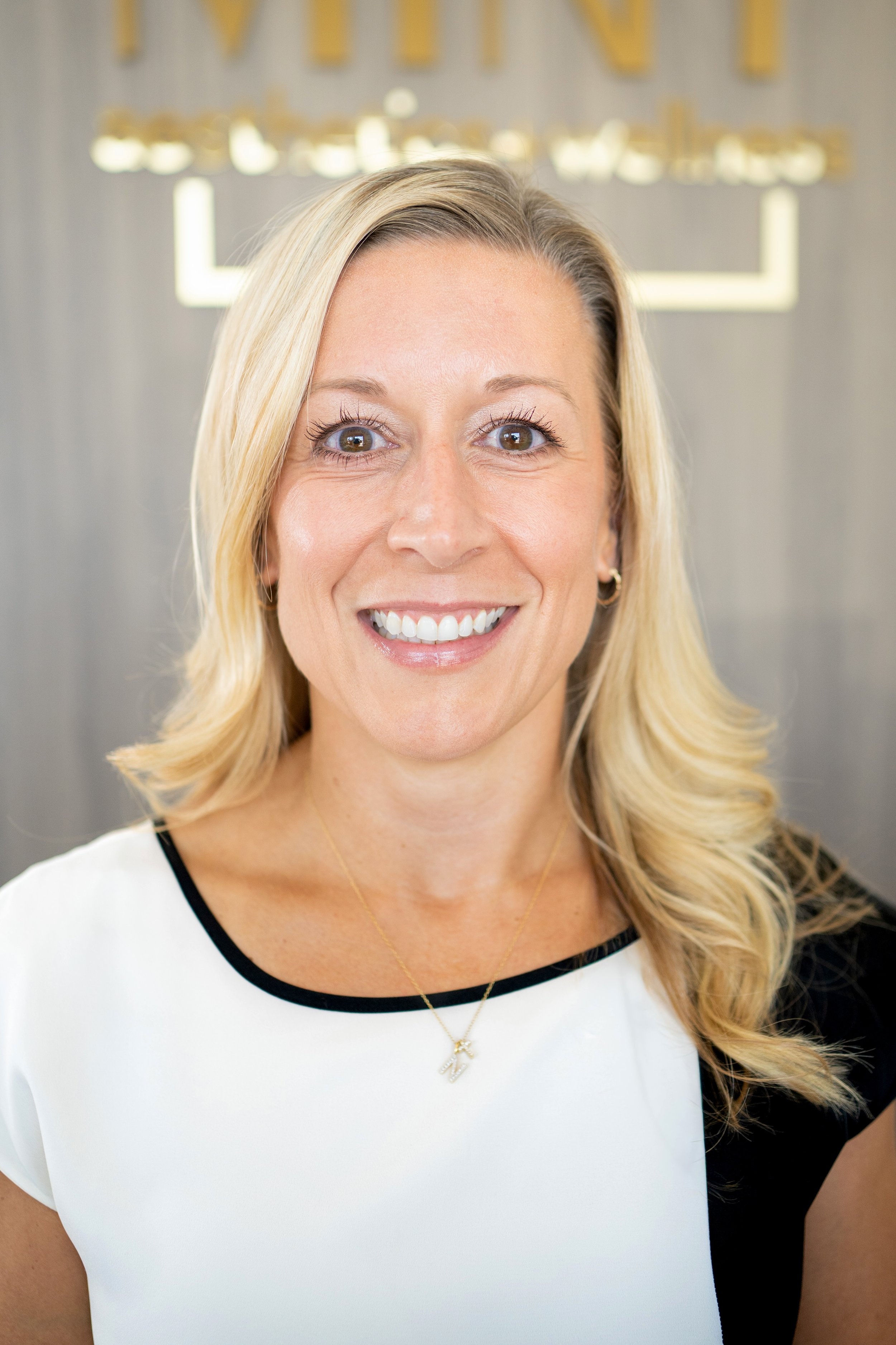 A woman with blonde hair smiling, wearing a navy blue jacket with 'MINT' and 'aesthetics & wellness' embroidered on it, looking at the camera against a plain, light-colored wall.
